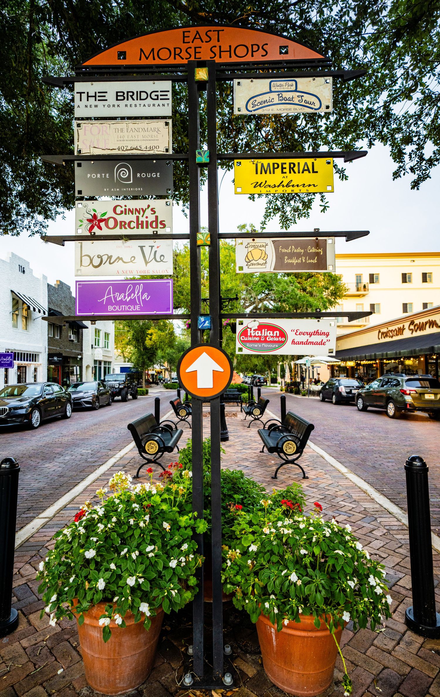 Shopping plaza with decorative planters and multiple business signs along a brick-paved walkway.