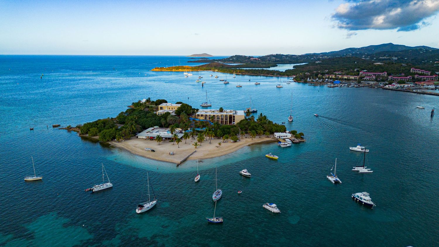 Aerial view of a lush tropical island with white sand beach, anchored sailboats, and turquoise Caribbean waters.