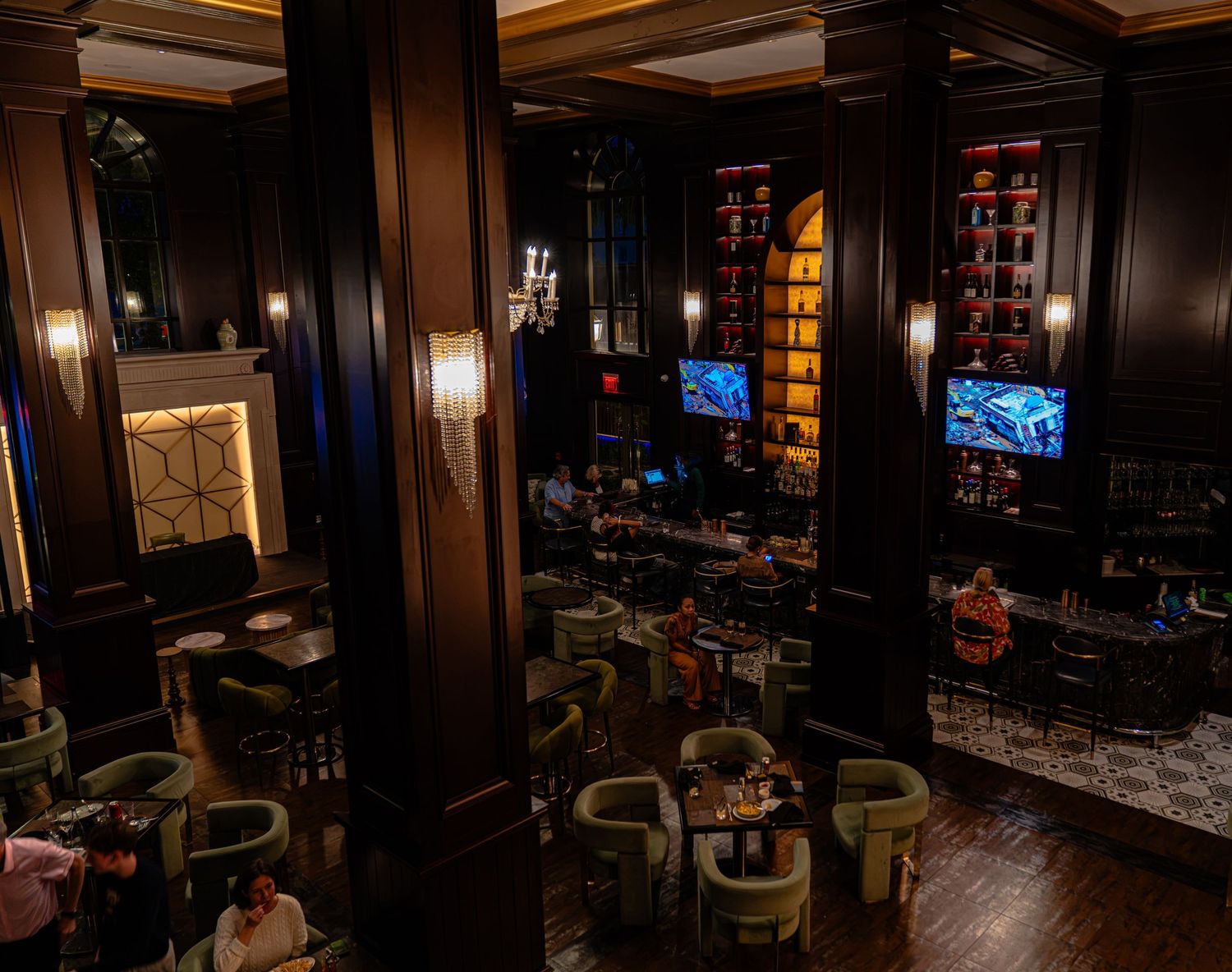 Elegant upscale bar lounge with dark wood paneling, green velvet chairs, glowing TVs, and ambient lighting.