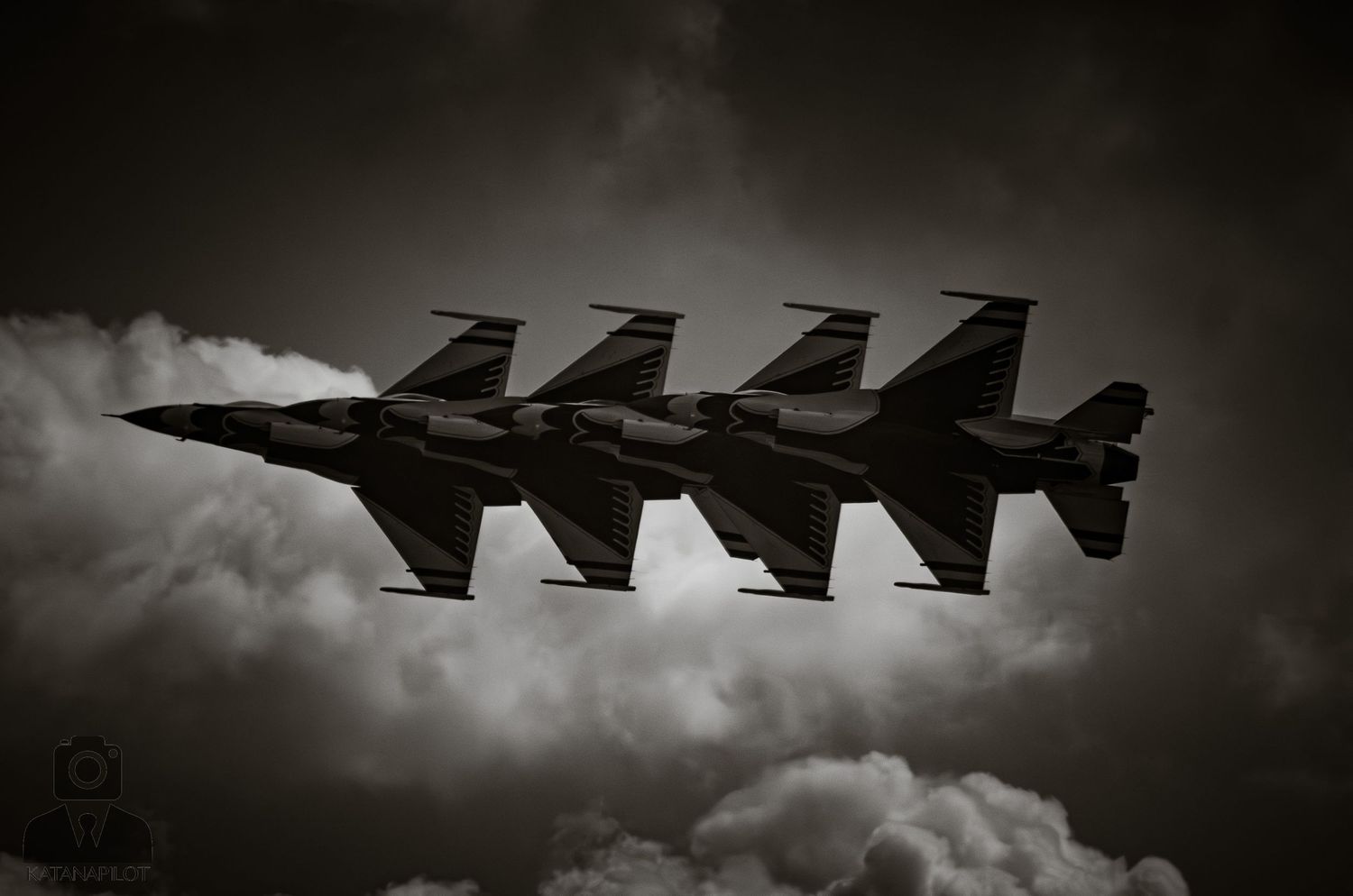 Four military jets fly in tight formation through dramatic cloudy skies in a striking black and white aerial photograph.
