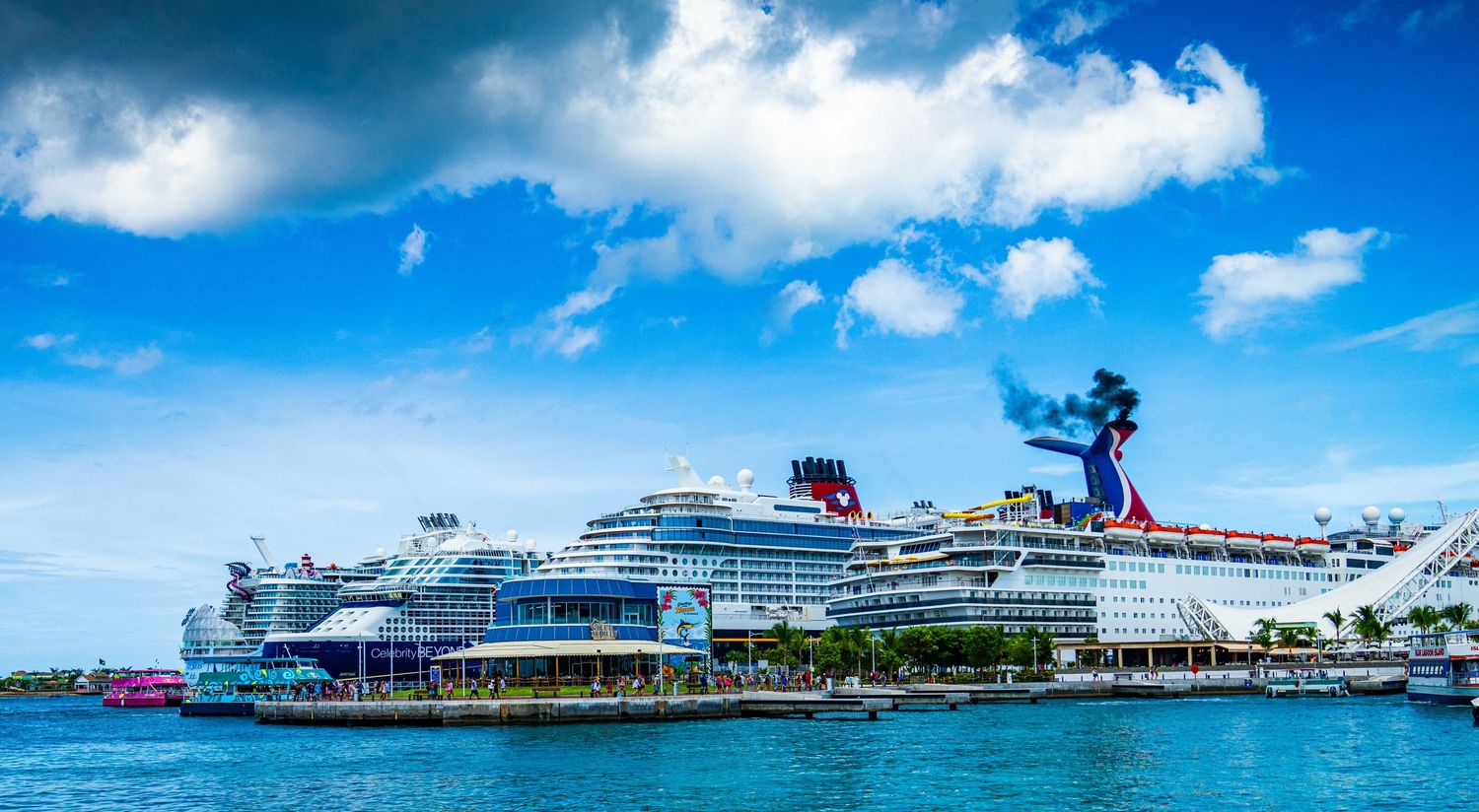 Large Disney cruise ship docked at a tropical port terminal under a bright blue sky with fluffy white clouds.