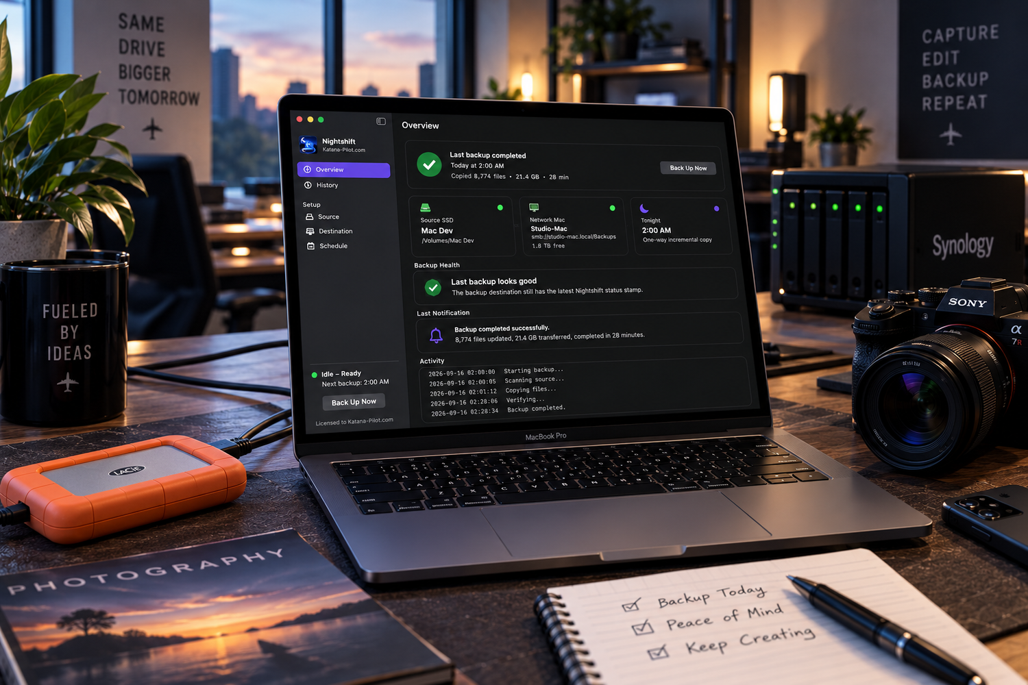A MacBook displaying Synology backup software on a desk with a LaCie drive, camera, and photography notebook at sunset.