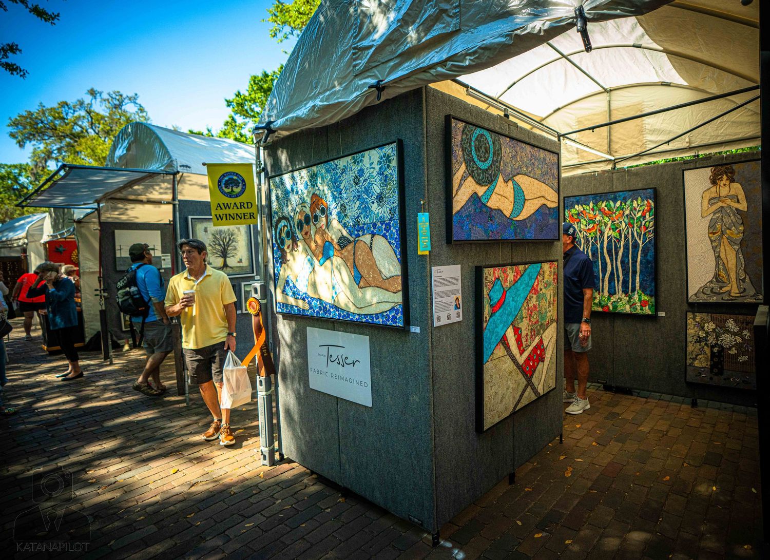 Art vendors display colorful paintings and artwork in outdoor market booths under canopies.