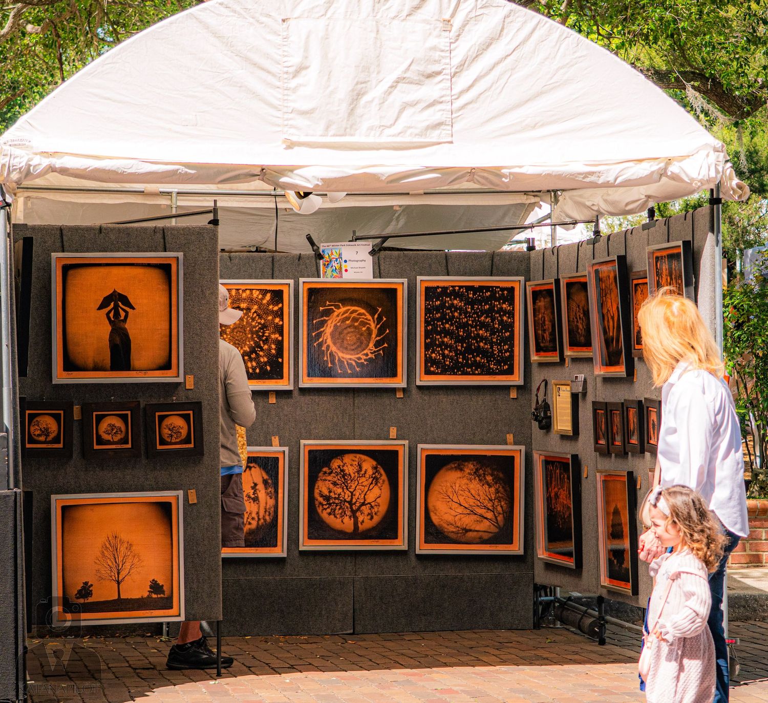 Art gallery booth displaying framed artwork on white walls under a tent canopy.
