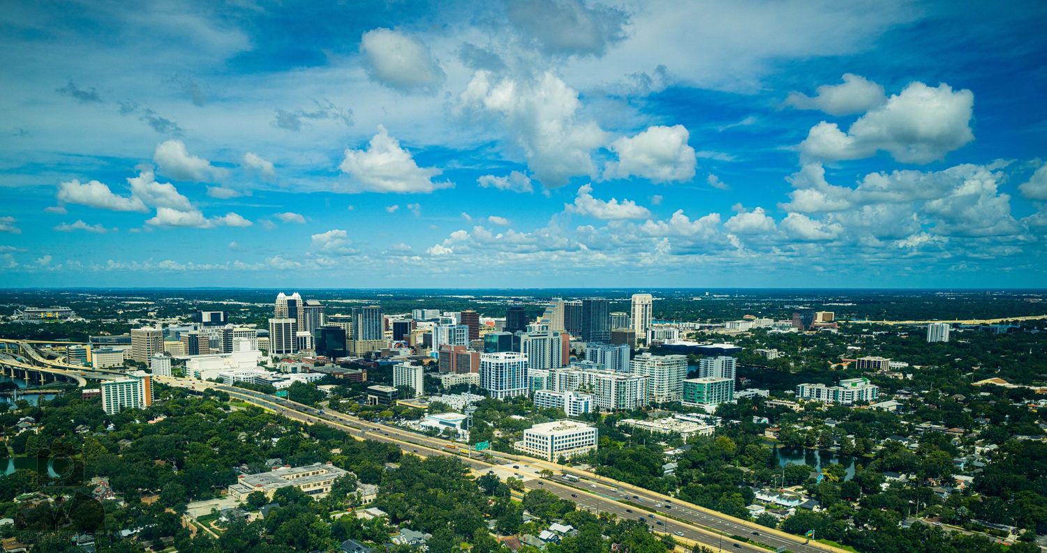 Panoramic cityscape view with modern buildings and infrastructure under puffy white clouds.
