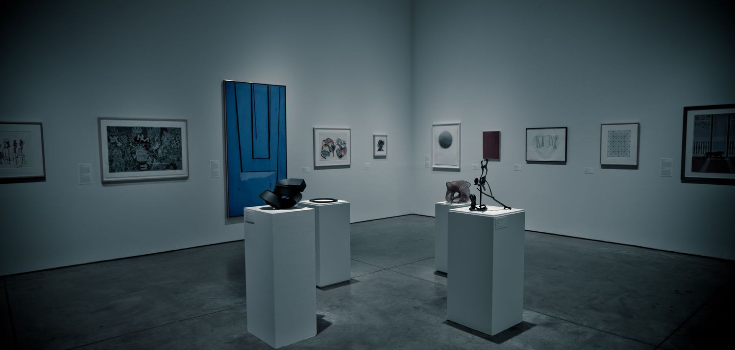 Art gallery interior with white pedestals displaying sculptures, framed artwork on walls, and a blue door accent.