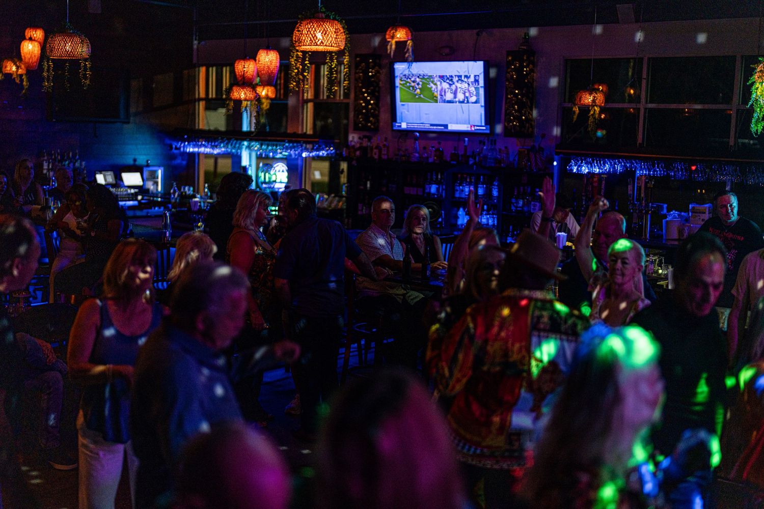 Crowded bar nightlife scene with colorful neon lights, dancing patrons, and a TV screen glowing in the background.