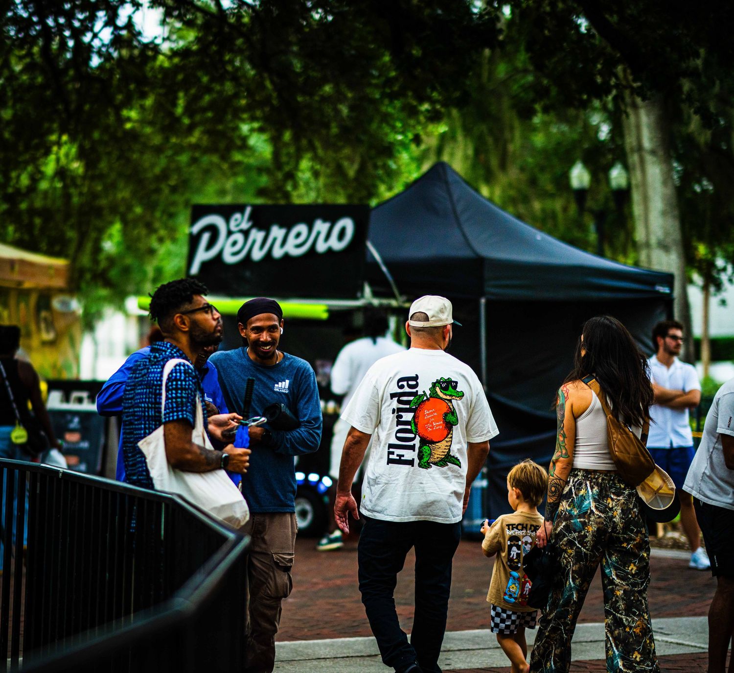 People walking at El Perrero outdoor food event in Florida, with vendors and lush green trees in background.