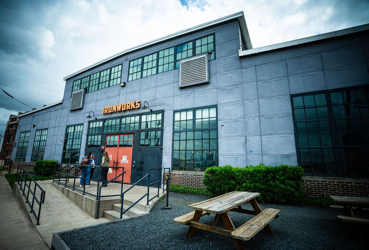Exterior of Ironworks brewery building with picnic table, large windows, and cloudy sky in an urban setting.