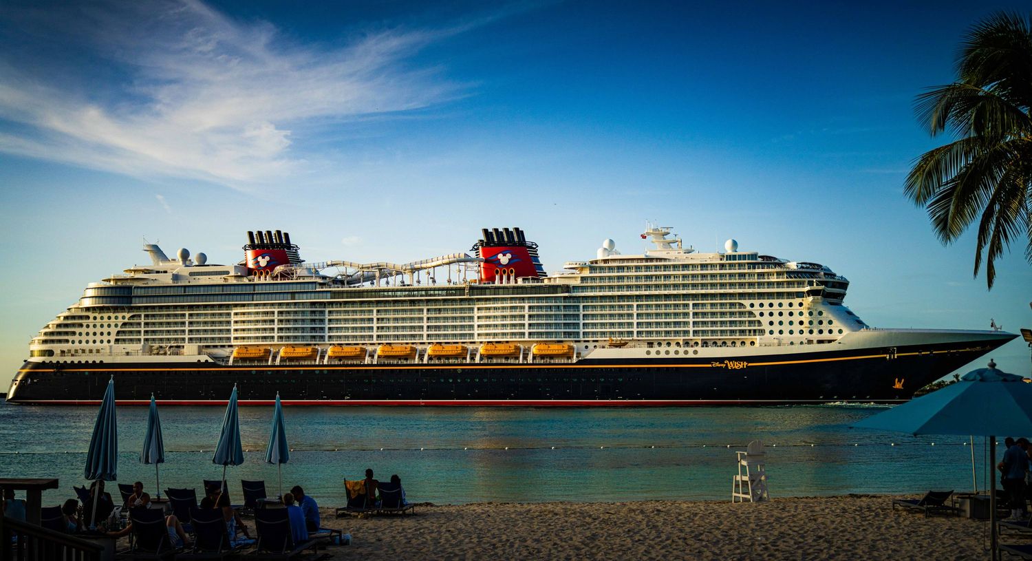 Disney cruise ship docked near a tropical beach with palm trees and beach chairs under a blue sky.