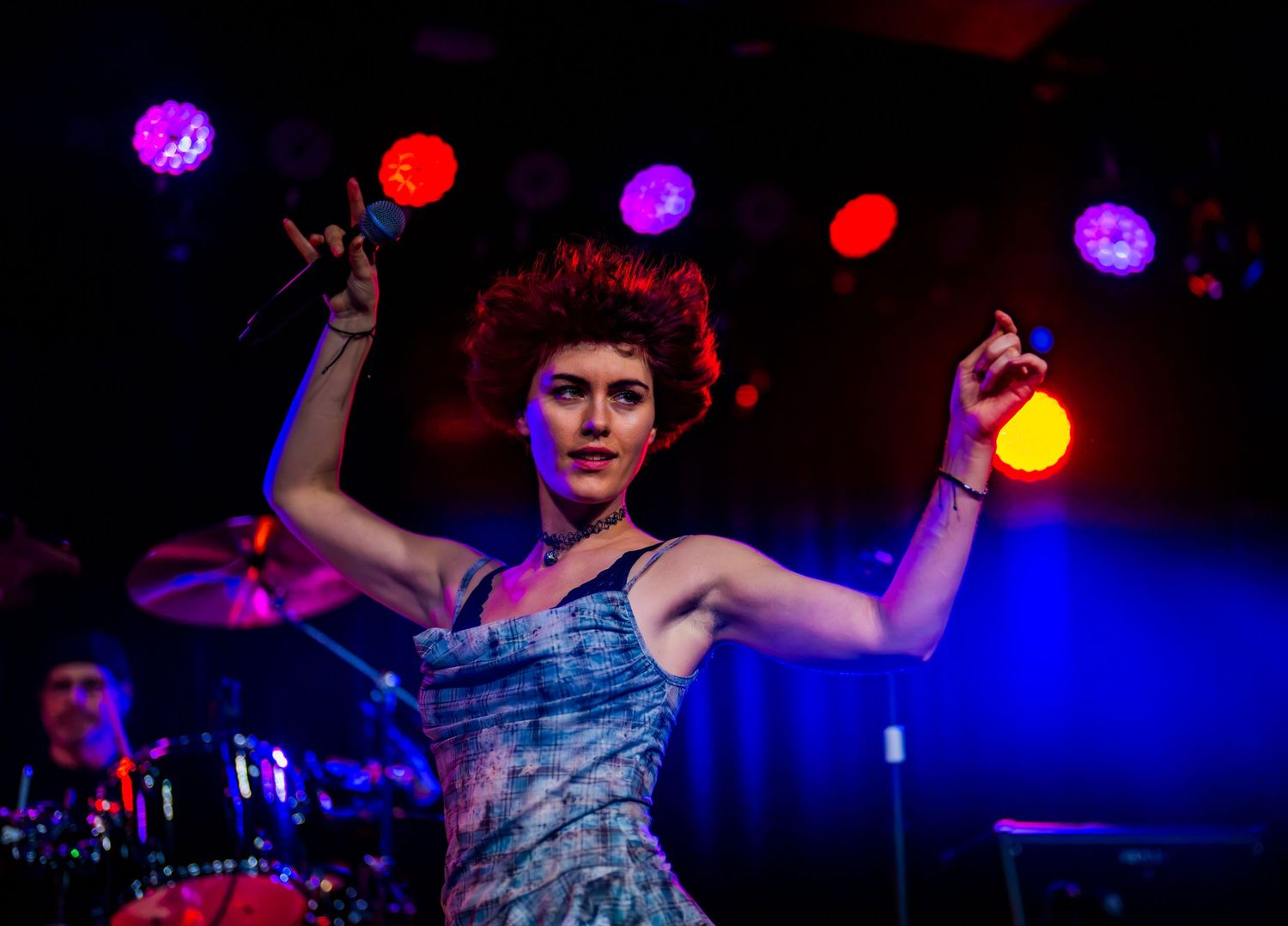 Female rock singer with red curly hair performs on stage, raising hands under colorful concert lights.