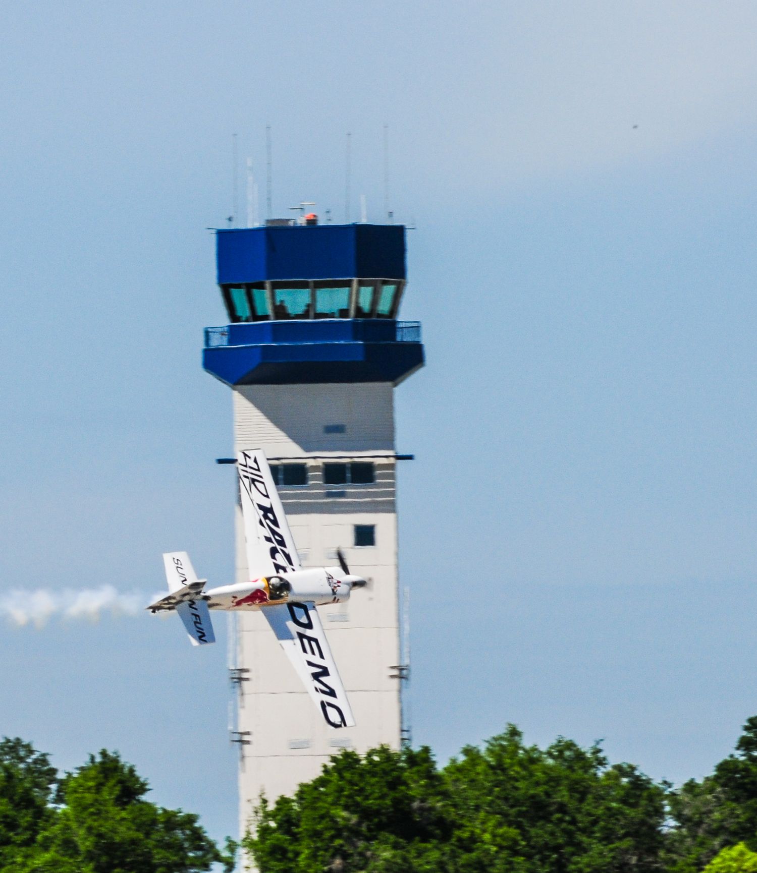 Small aircraft flying past a white air traffic control tower against blue sky.