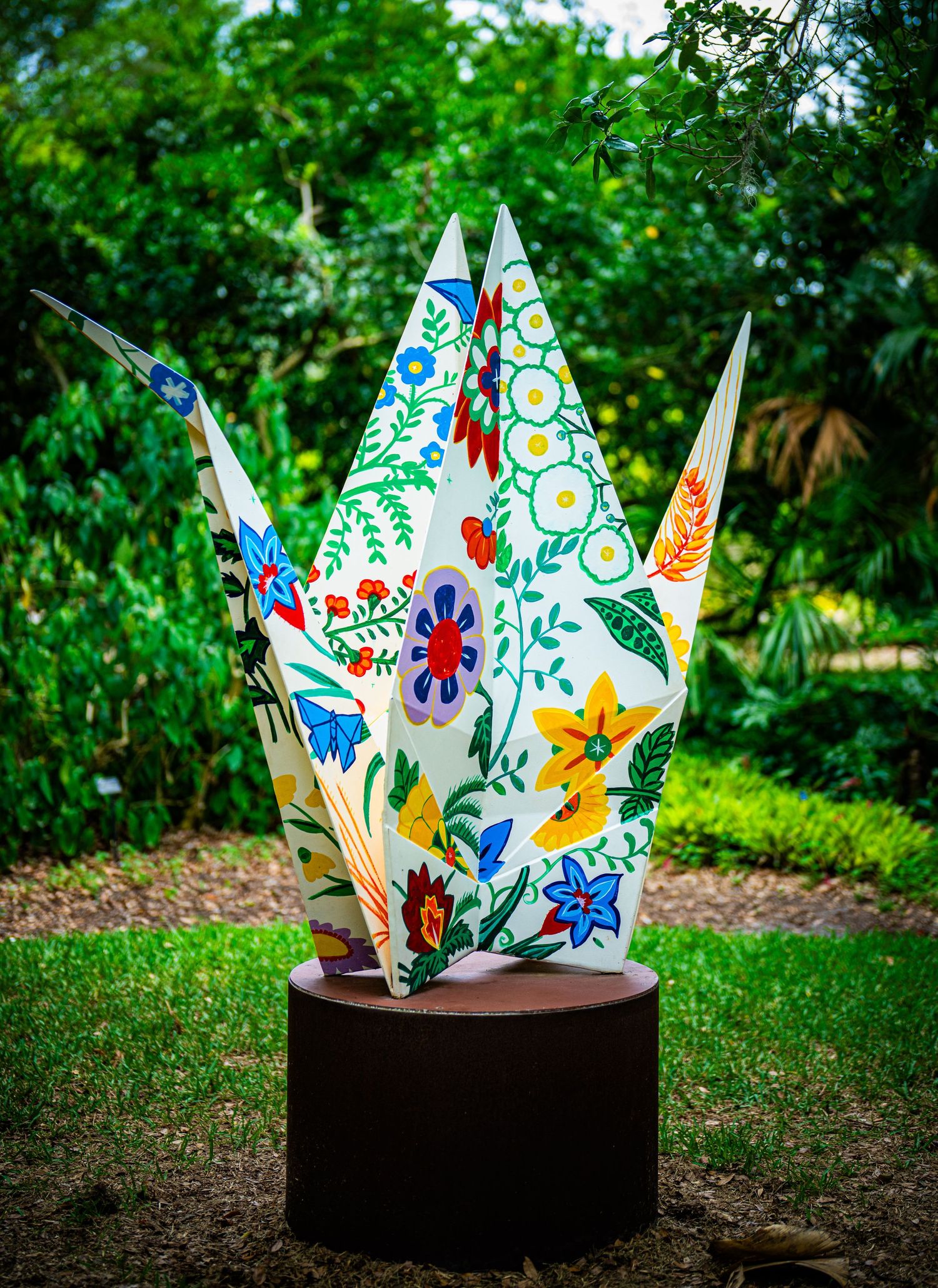 Colorful floral origami crown with painted designs displayed on dark pedestal outdoors.