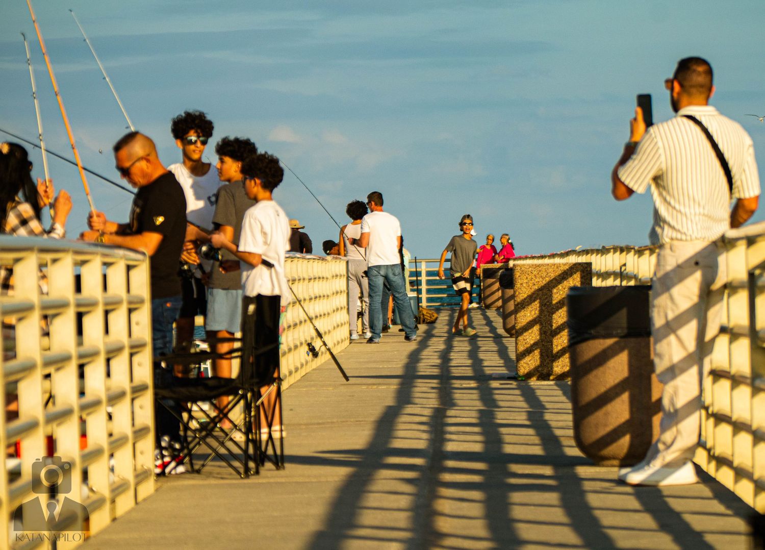 People enjoying a sunny afternoon on a long wooden pier with dramatic shadows cast by the railing.