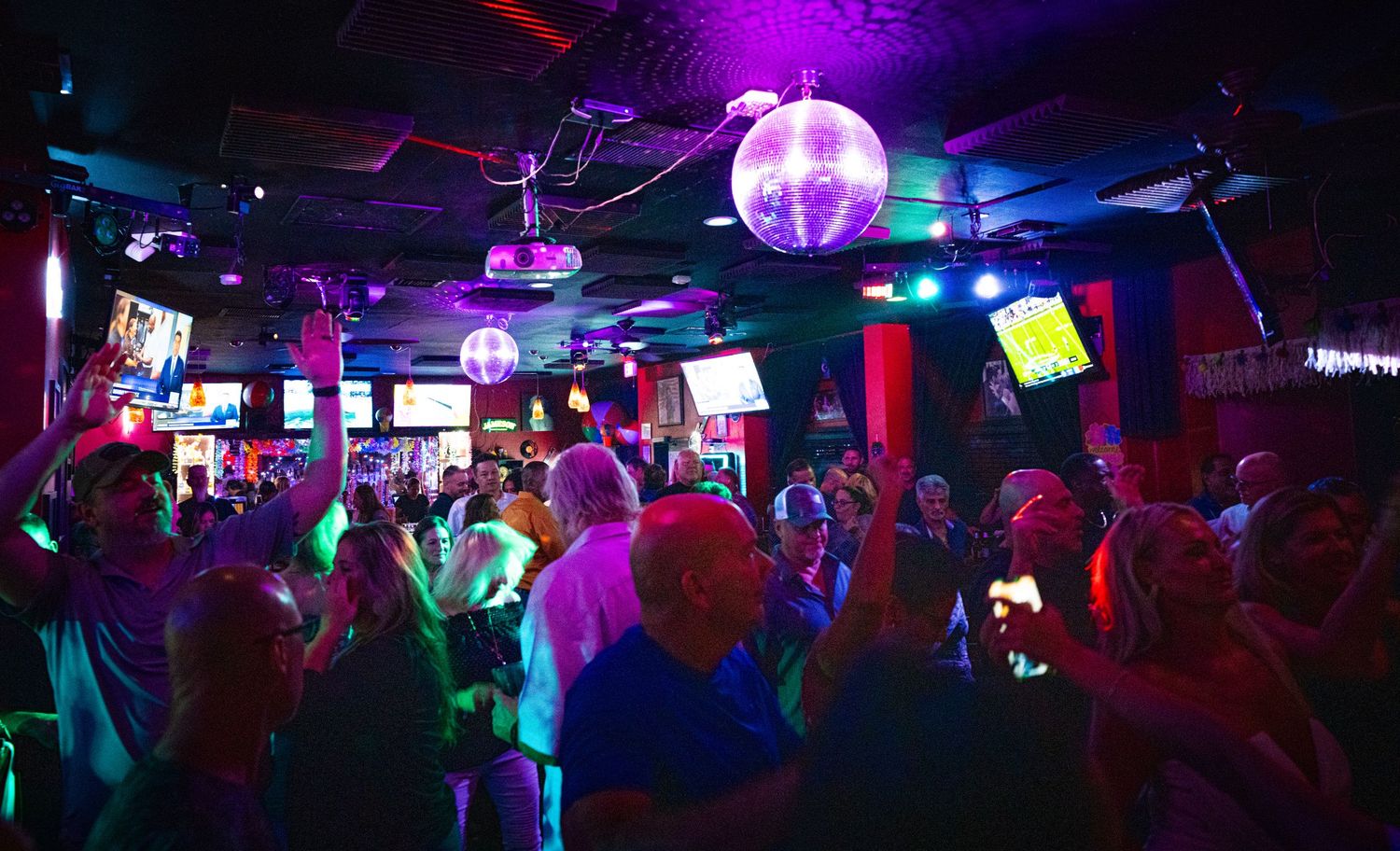 Colorful disco lights illuminate a crowded dance floor with a glowing purple disco ball overhead.