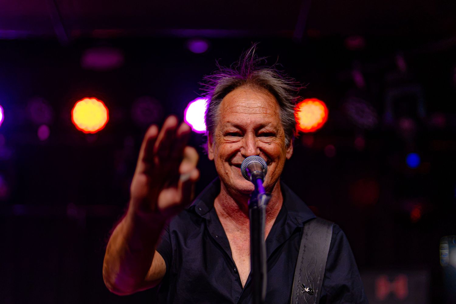 Male rock musician smiling and waving at crowd while performing on stage with colorful concert lights.
