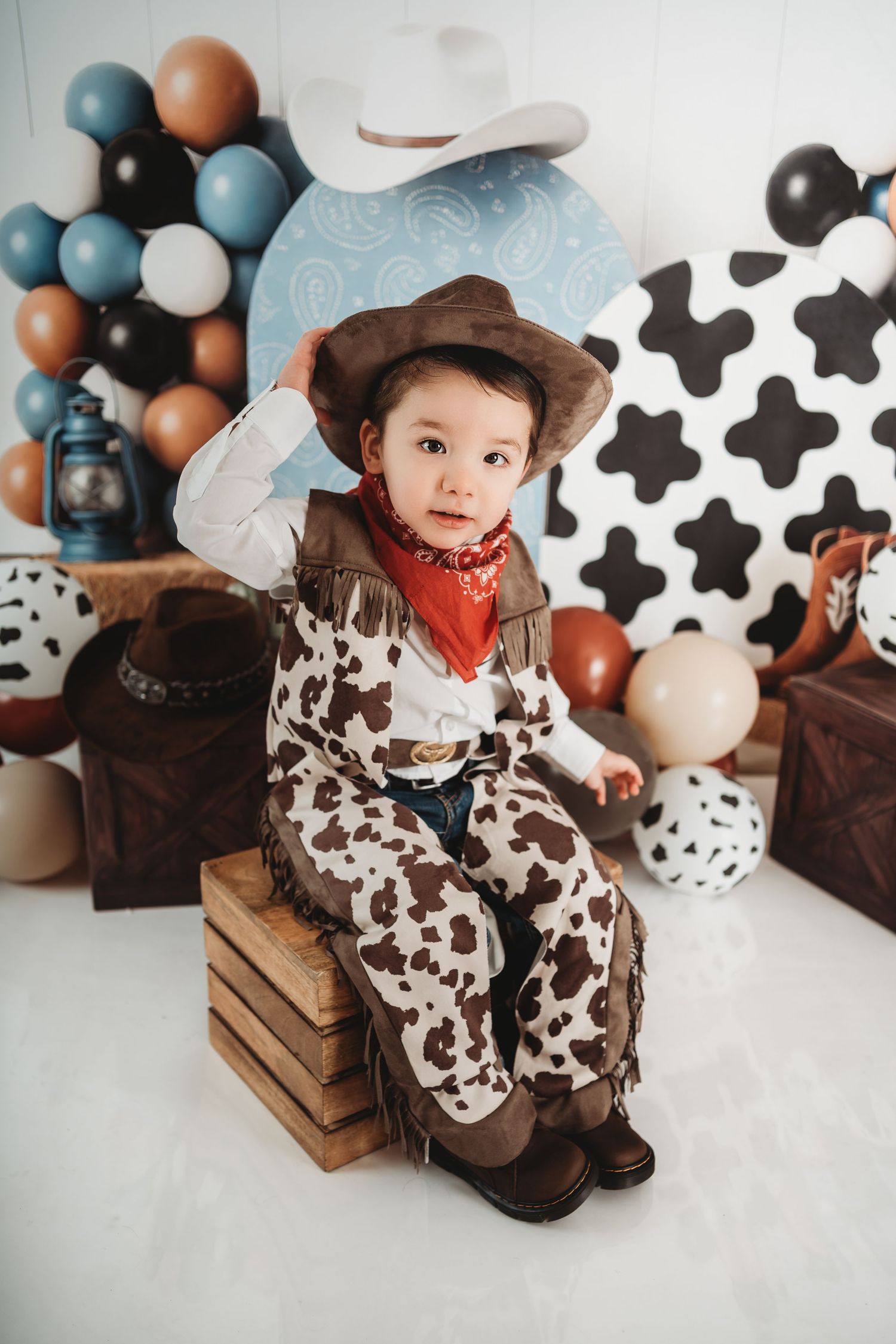 Toddler in cowboy costume with hat, vest, and bandana sits on wooden crates at a Western-themed photo shoot.