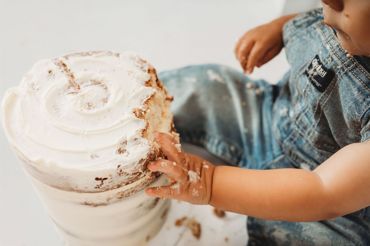 Toddler in denim overalls smashing a white frosted cake with their hands during a cake smash photoshoot.