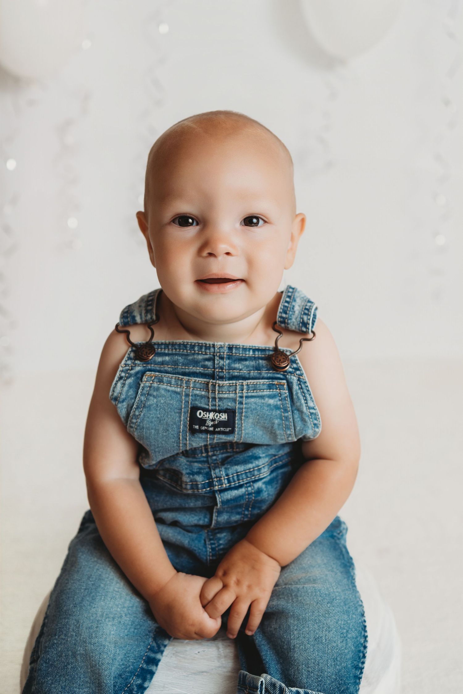Smiling baby in OshKosh denim overalls sitting upright against a white background with balloons.