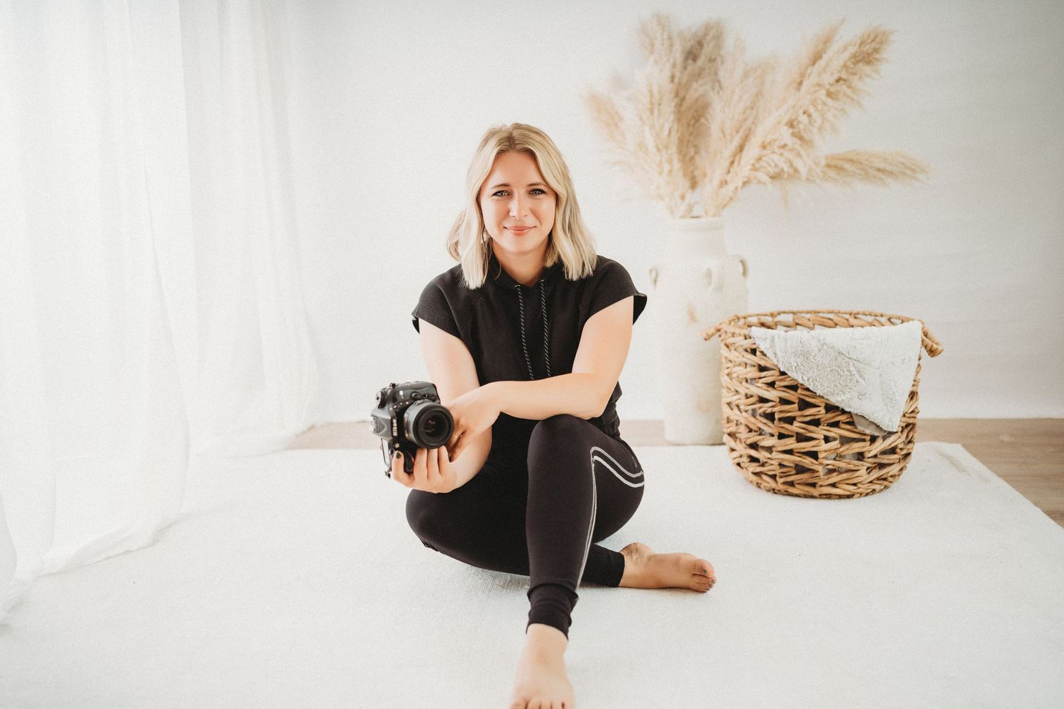 Blonde woman in black outfit sitting on white floor holding camera in bright minimalist studio with pampas grass decor.