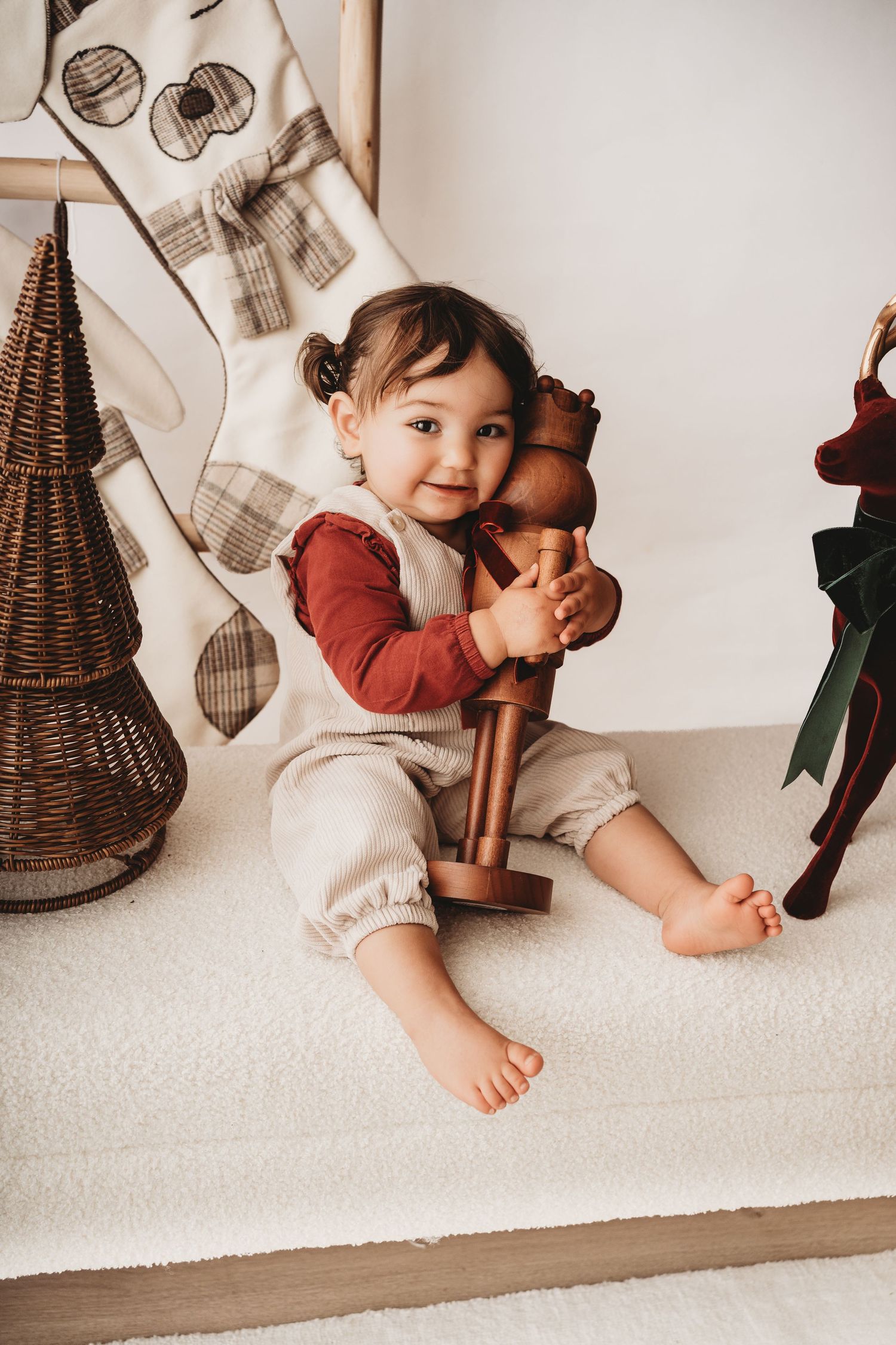 A smiling toddler girl hugs a wooden nutcracker toy while sitting on a white rug with holiday decorations nearby.