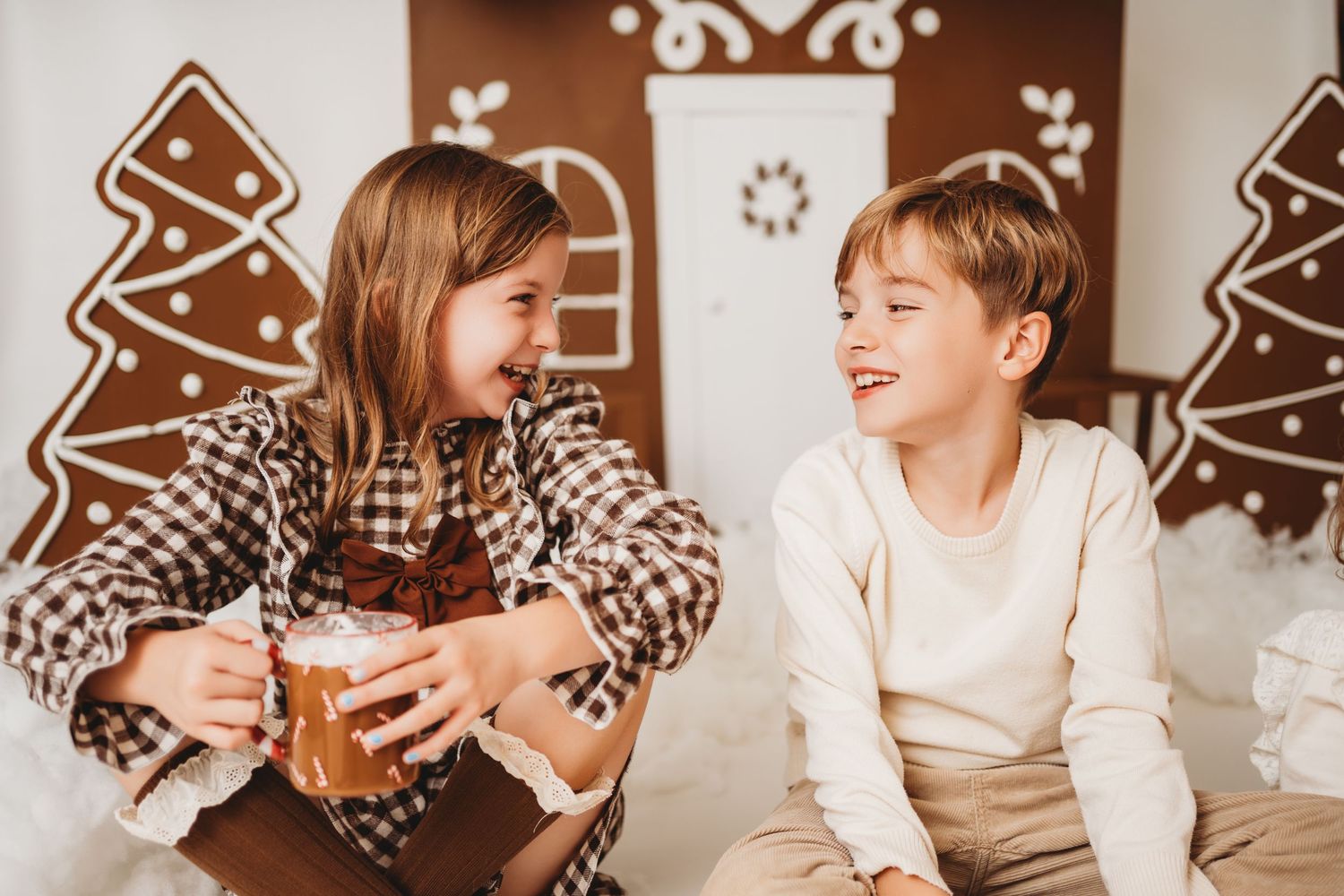Two happy children sharing hot cocoa in front of a festive gingerbread house Christmas backdrop.