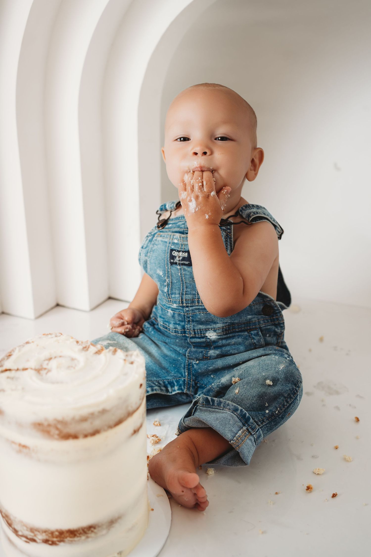 A baby in denim overalls sits on a white floor, eating cake with their hands beside a white frosted smash cake.