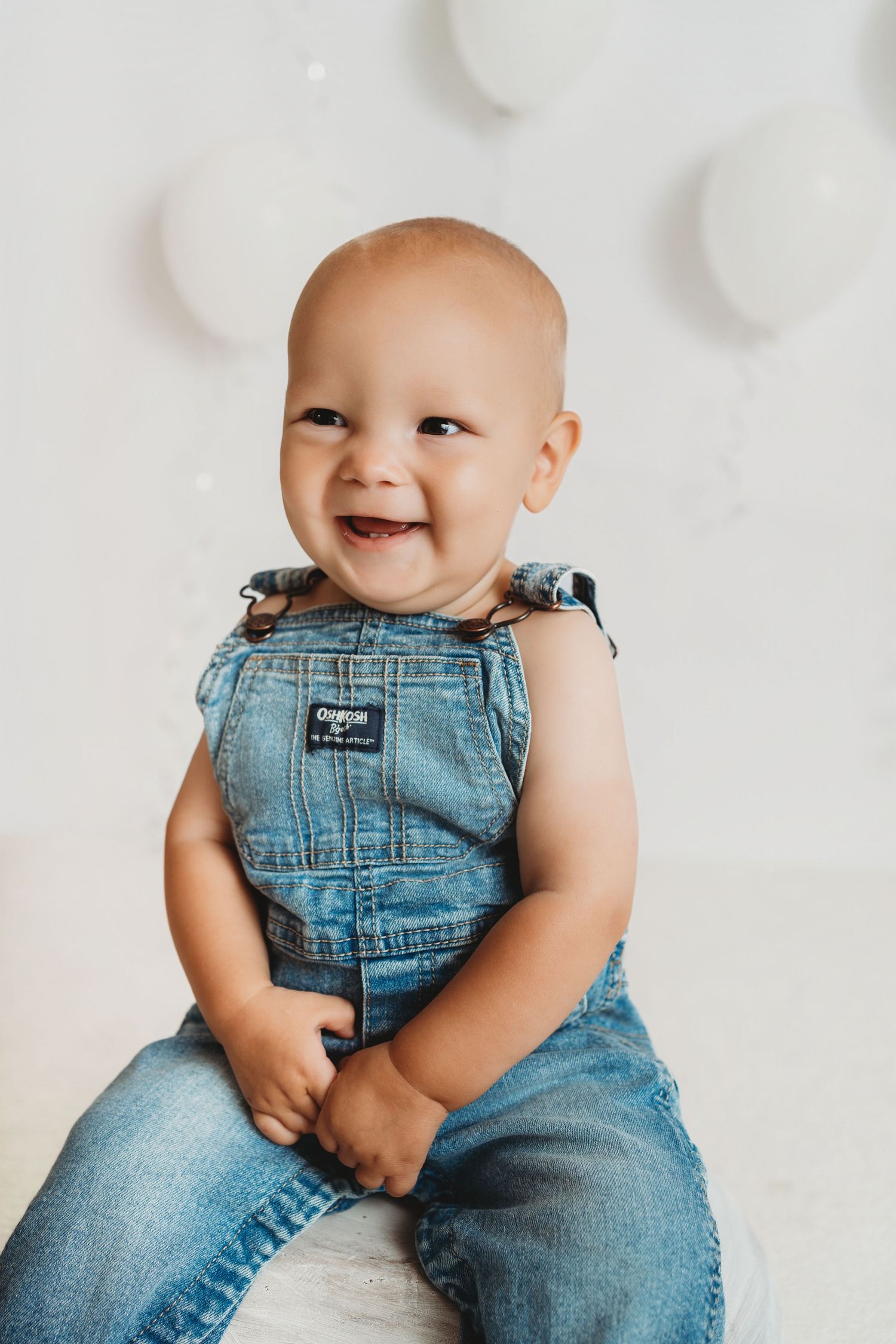 Smiling baby in denim overalls sitting against a white background with balloons.
