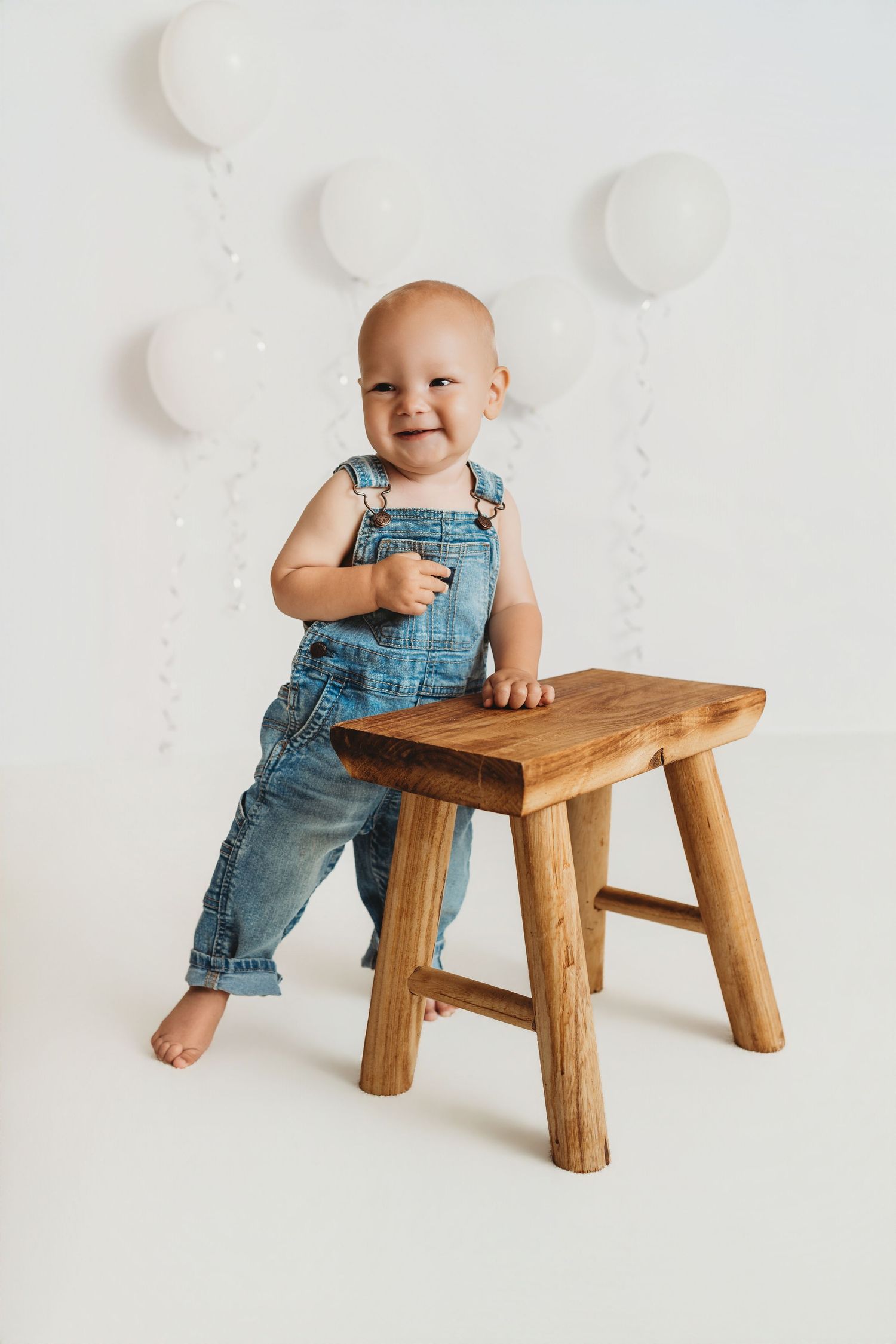 Smiling baby in denim overalls standing next to a wooden stool with white balloons in the background.