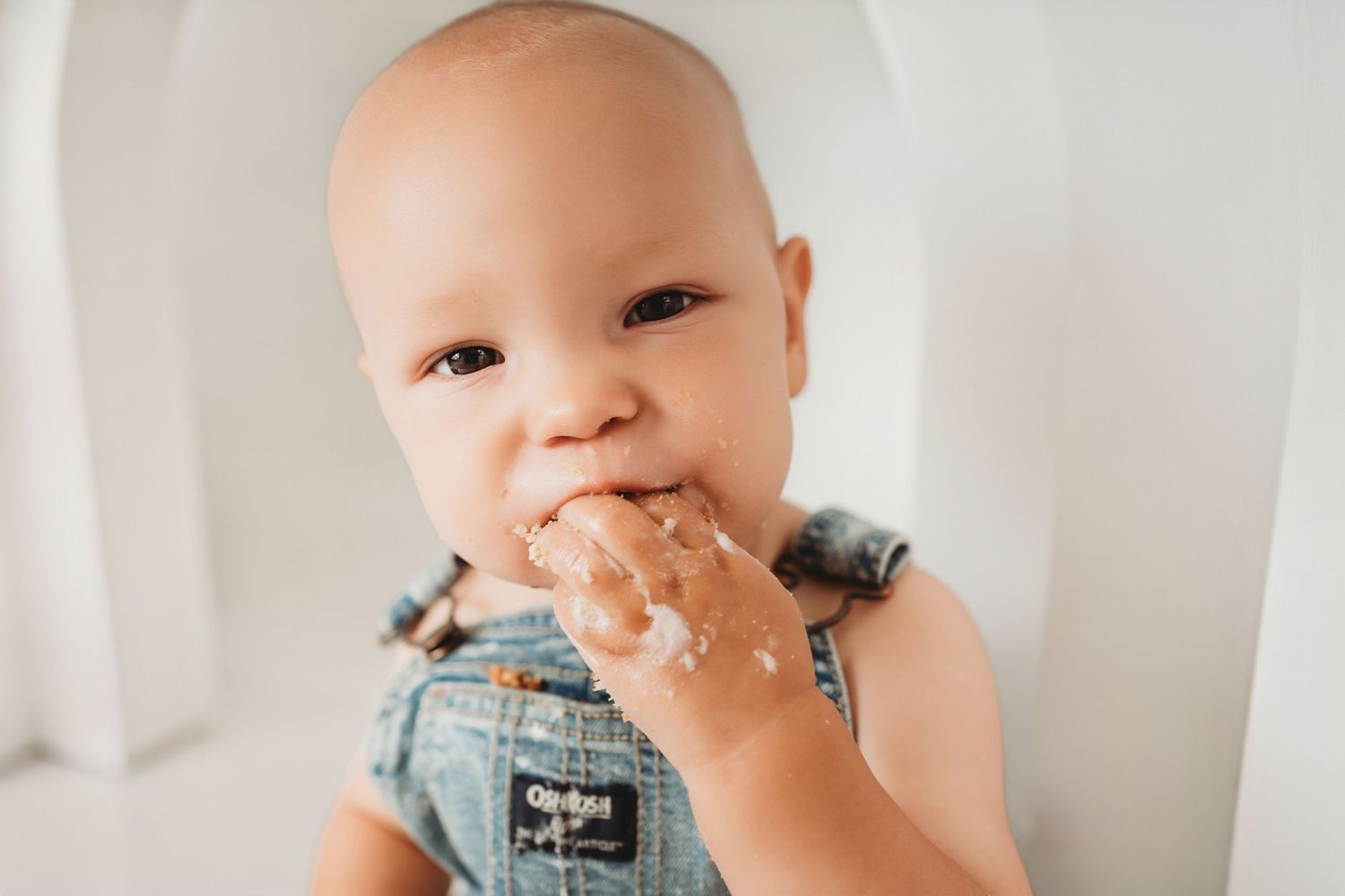 Cute baby in OshKosh denim overalls eating cake with messy hands in a white high chair.