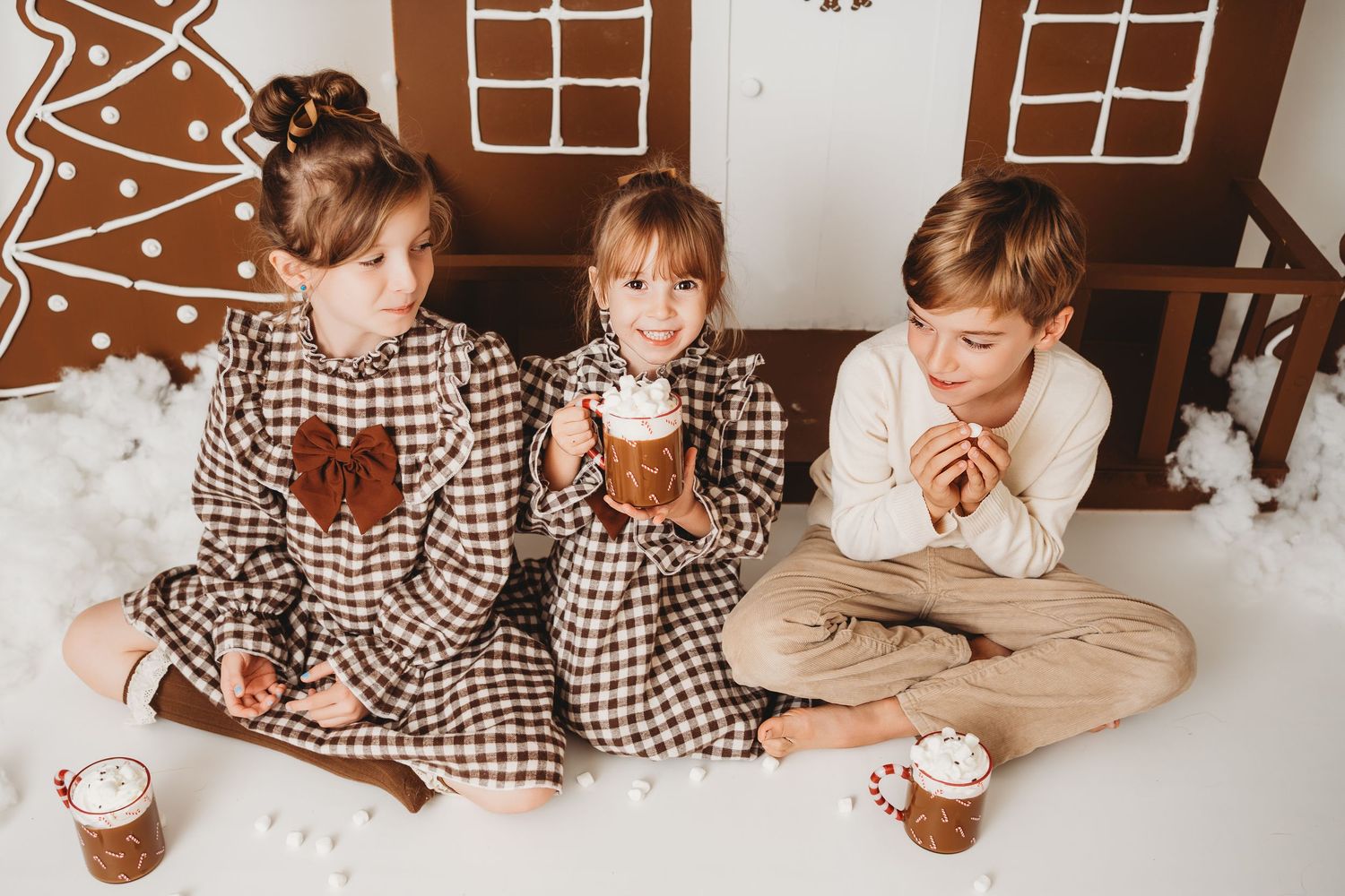 Three children in festive pajamas sit together enjoying hot cocoa near a gingerbread house backdrop during Christmas.