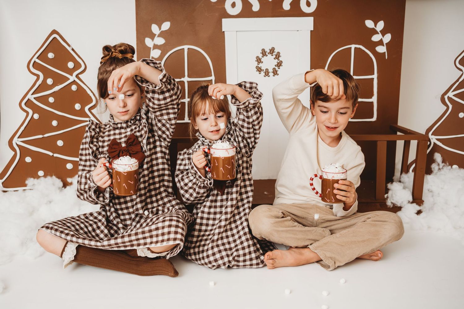 Three children in plaid pajamas sit together holding mugs of hot cocoa with marshmallows near a gingerbread house backdrop.