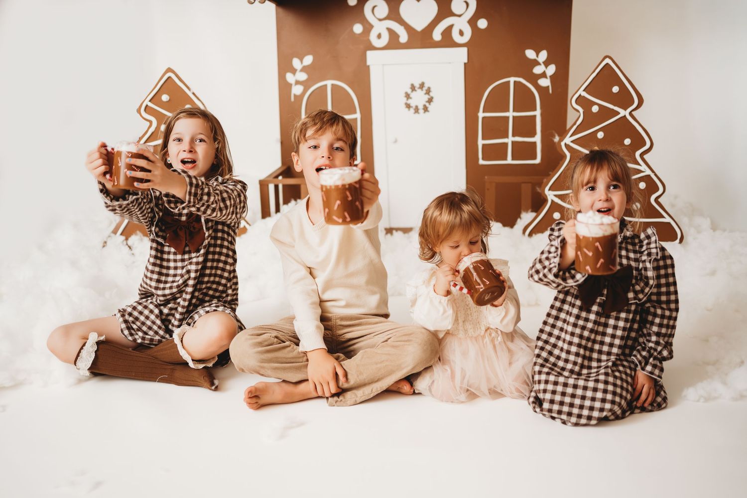 Four children sit together holding gingerbread cookies and mugs near a decorative cardboard gingerbread house backdrop.