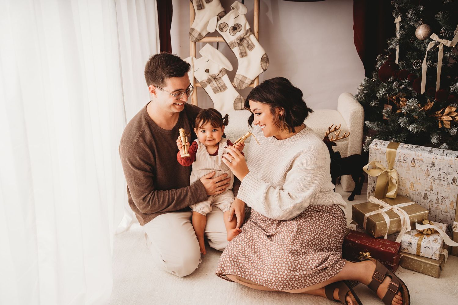 Family of three celebrating Christmas together near a decorated tree with gifts, sharing a joyful moment with their toddler.