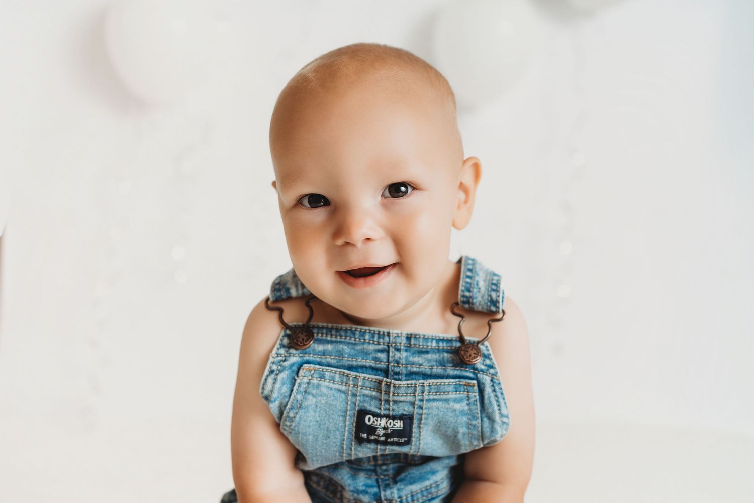 Smiling baby in denim OshKosh overalls posing for a portrait against a soft white background.