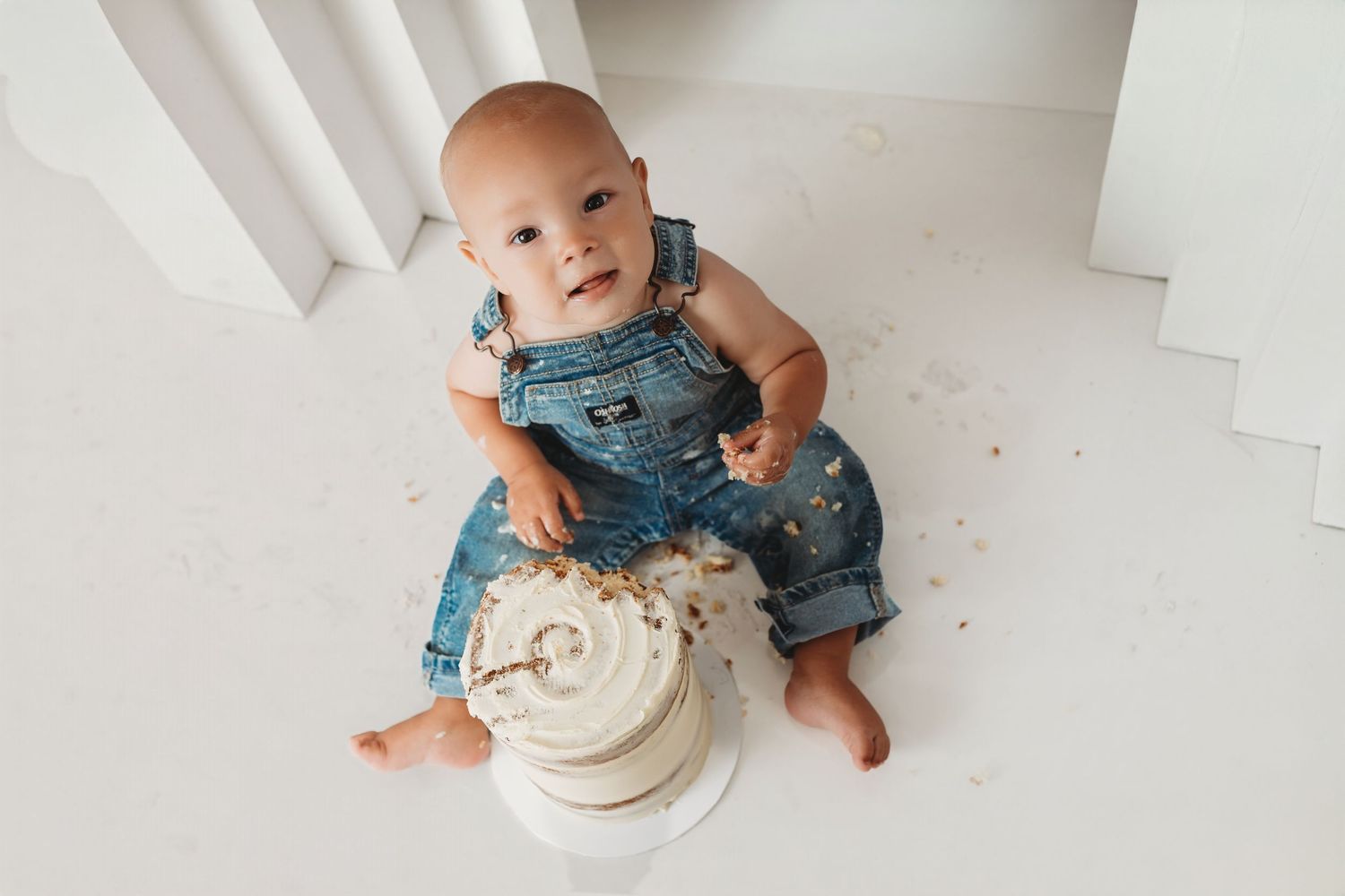 Baby in denim overalls sitting on floor with smashed first birthday cake during cake smash photo session.