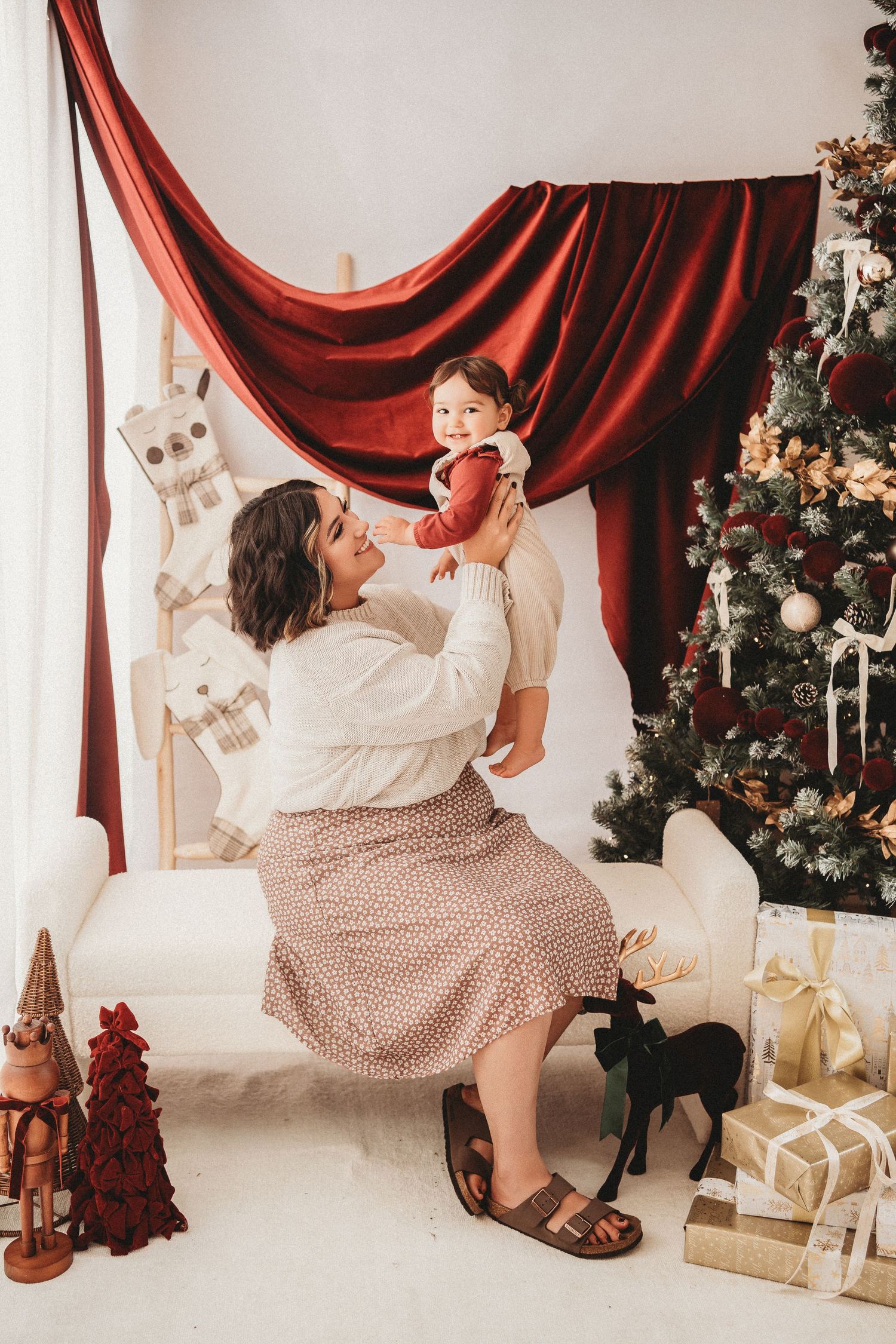 Mother lifting baby near Christmas tree with red drape, gifts, and holiday decorations in festive white room.