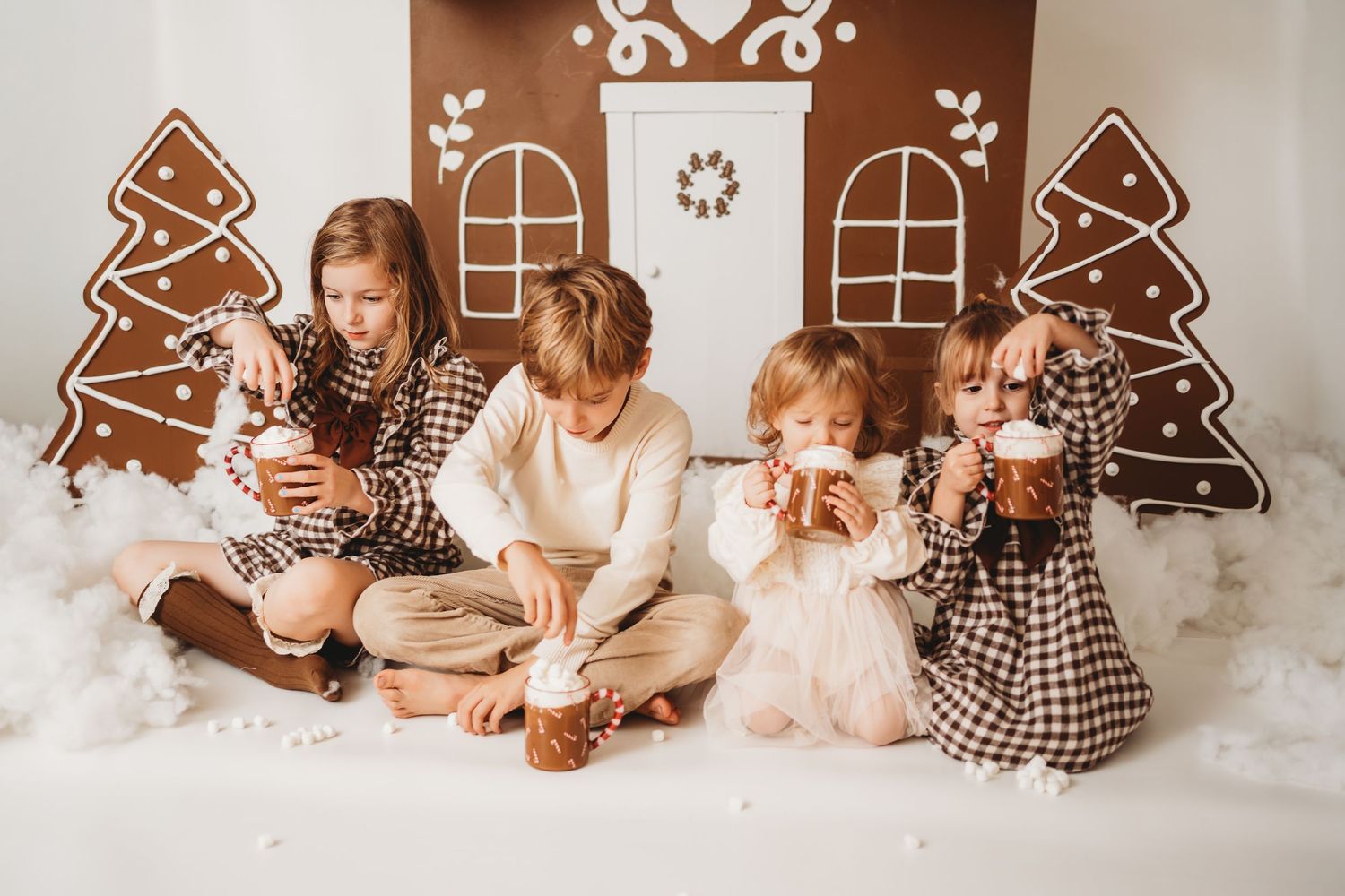 Four children sit together in a festive gingerbread house Christmas photo set, wearing matching plaid outfits.