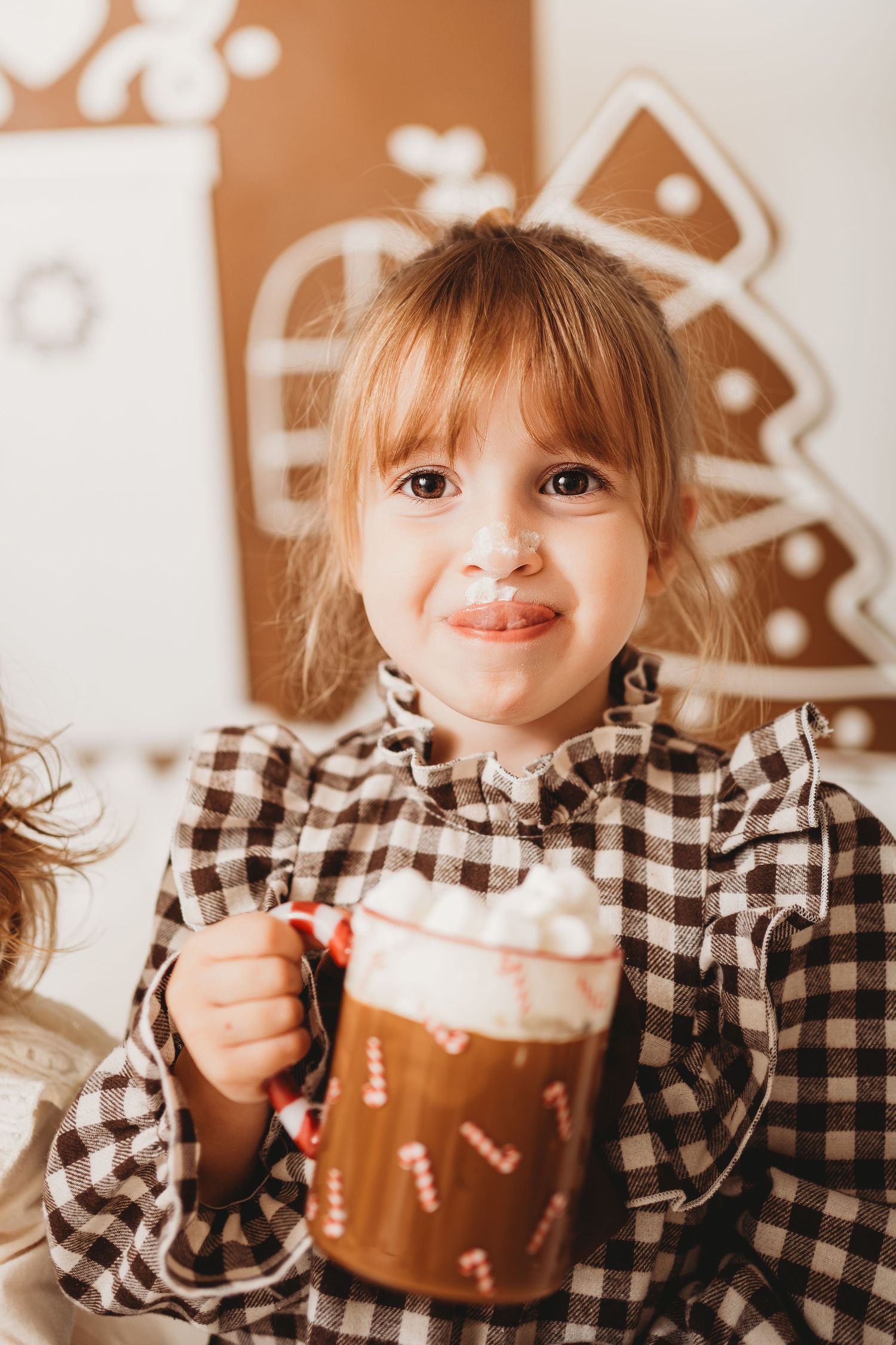 Young girl in plaid dress holding a festive mug with marshmallows, smiling in front of gingerbread Christmas decorations.
