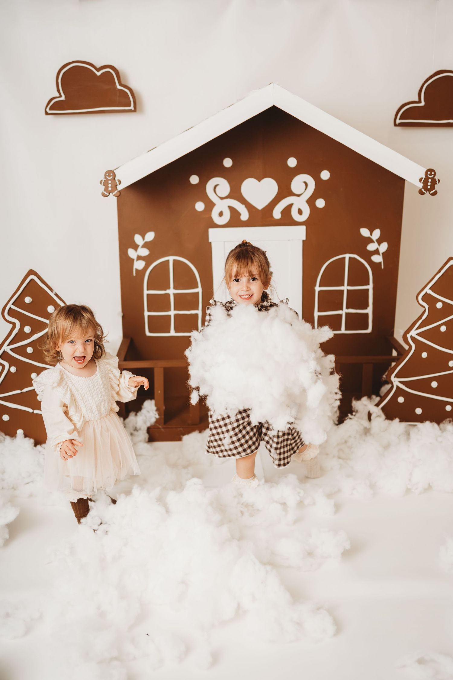 Two little girls playing with fluffy white snow in front of a decorative gingerbread house Christmas backdrop.