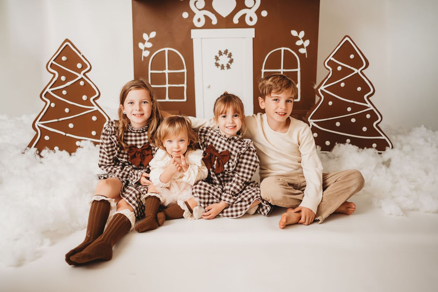 Four children pose together in front of a festive gingerbread house backdrop wearing holiday plaid outfits.