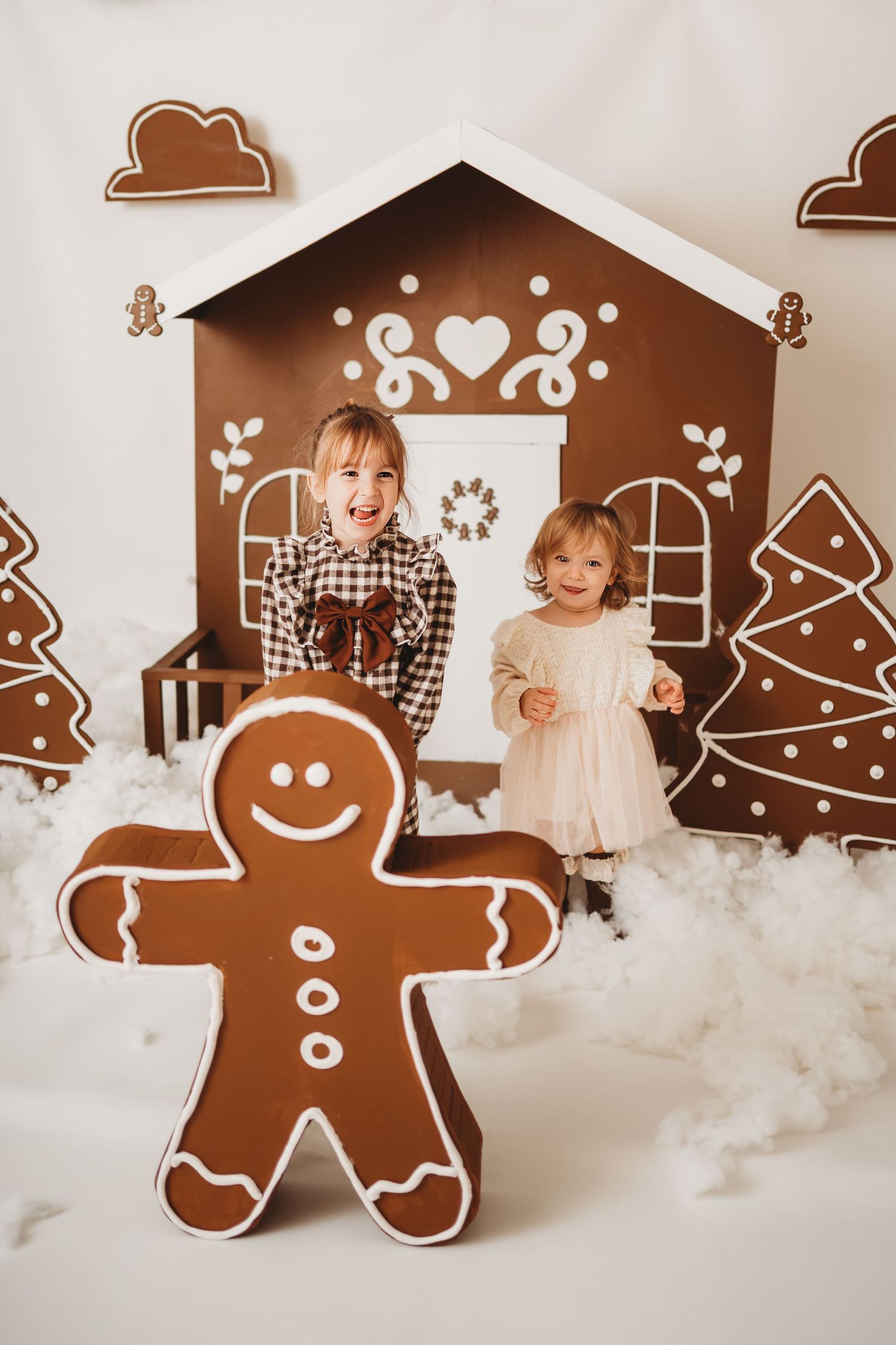 Two children posing with giant gingerbread man decoration in front of festive gingerbread house Christmas backdrop.