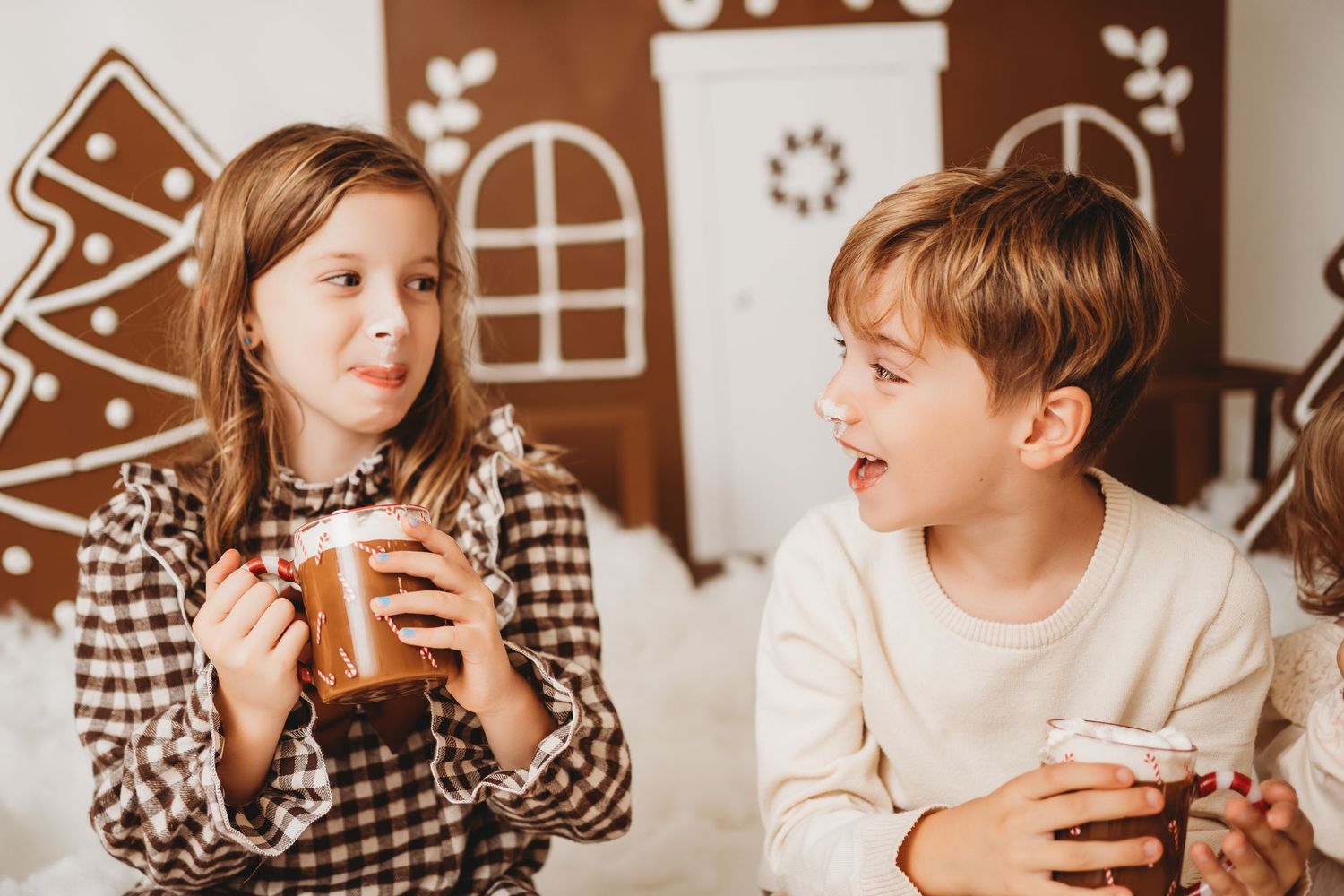 Two children enjoying hot cocoa mugs in front of festive gingerbread house decorations during Christmas holiday.
