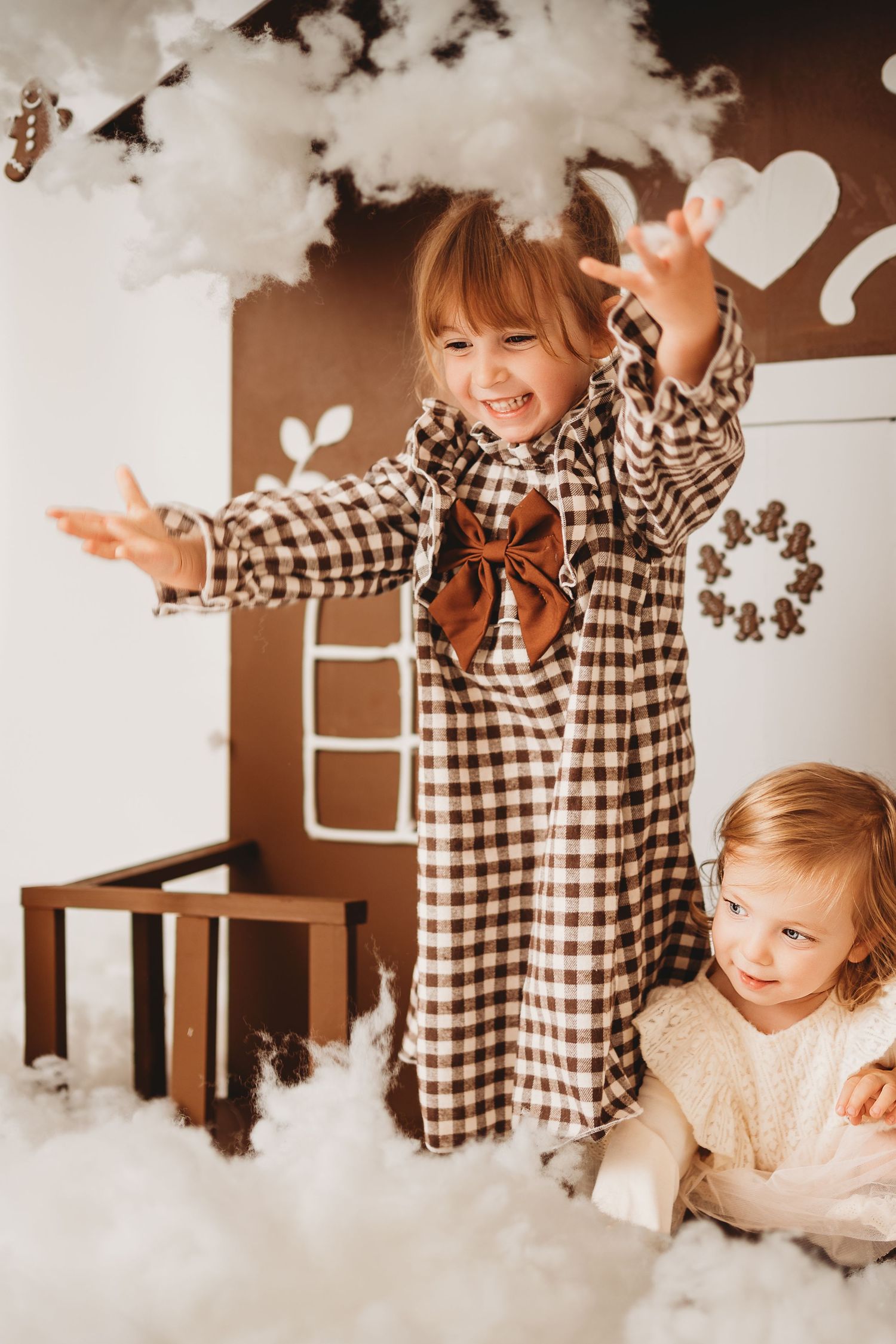 Two young girls playing in a festive Christmas decorated room with cotton snow clouds and gingerbread house backdrop.