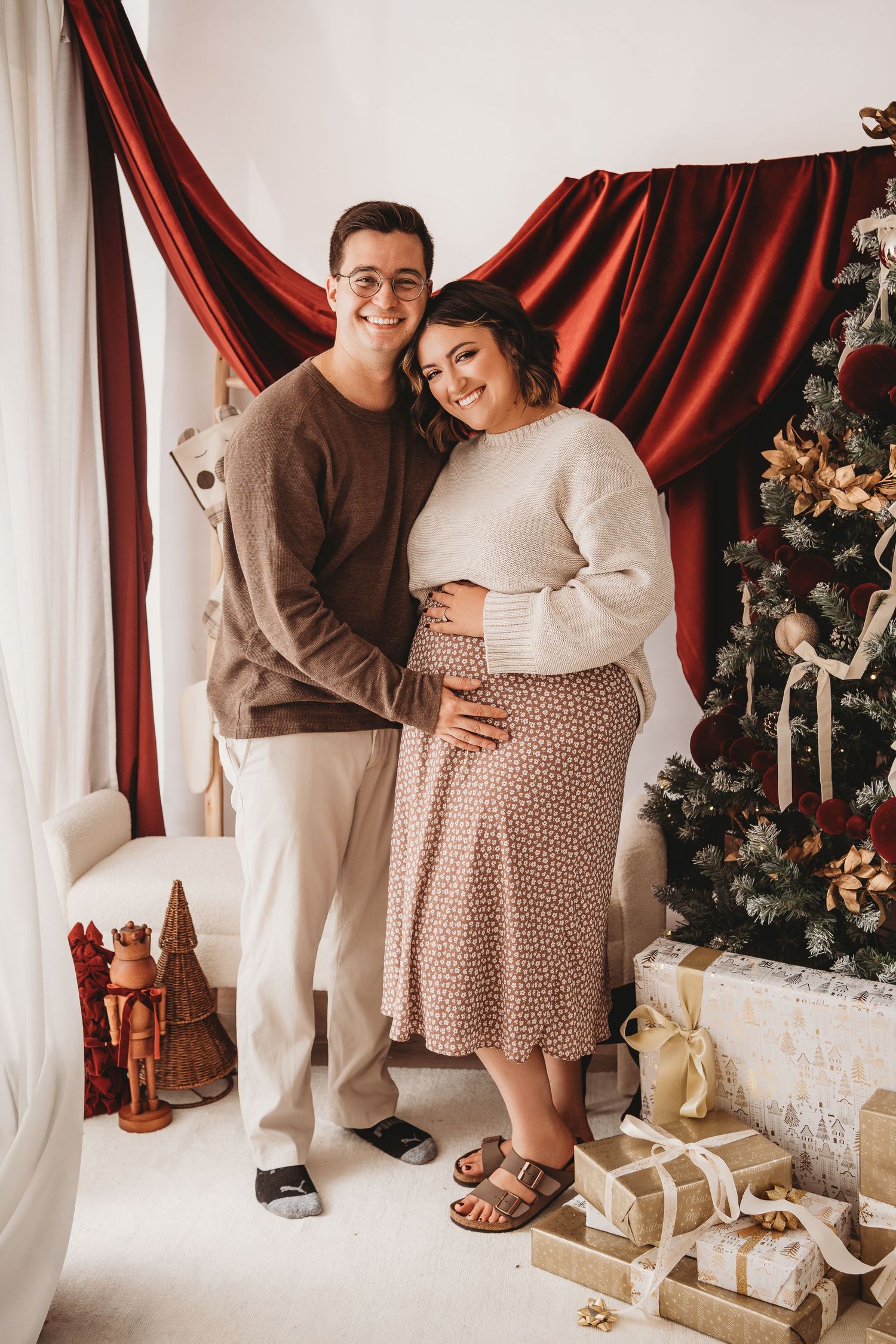 Happy couple poses by Christmas tree with gifts, woman in cream sweater and floral skirt holding baby bump.