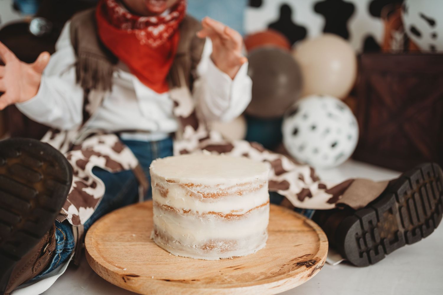 Baby in cowboy outfit sits behind a rustic naked cake on a wood slice at a western-themed smash cake photo shoot.