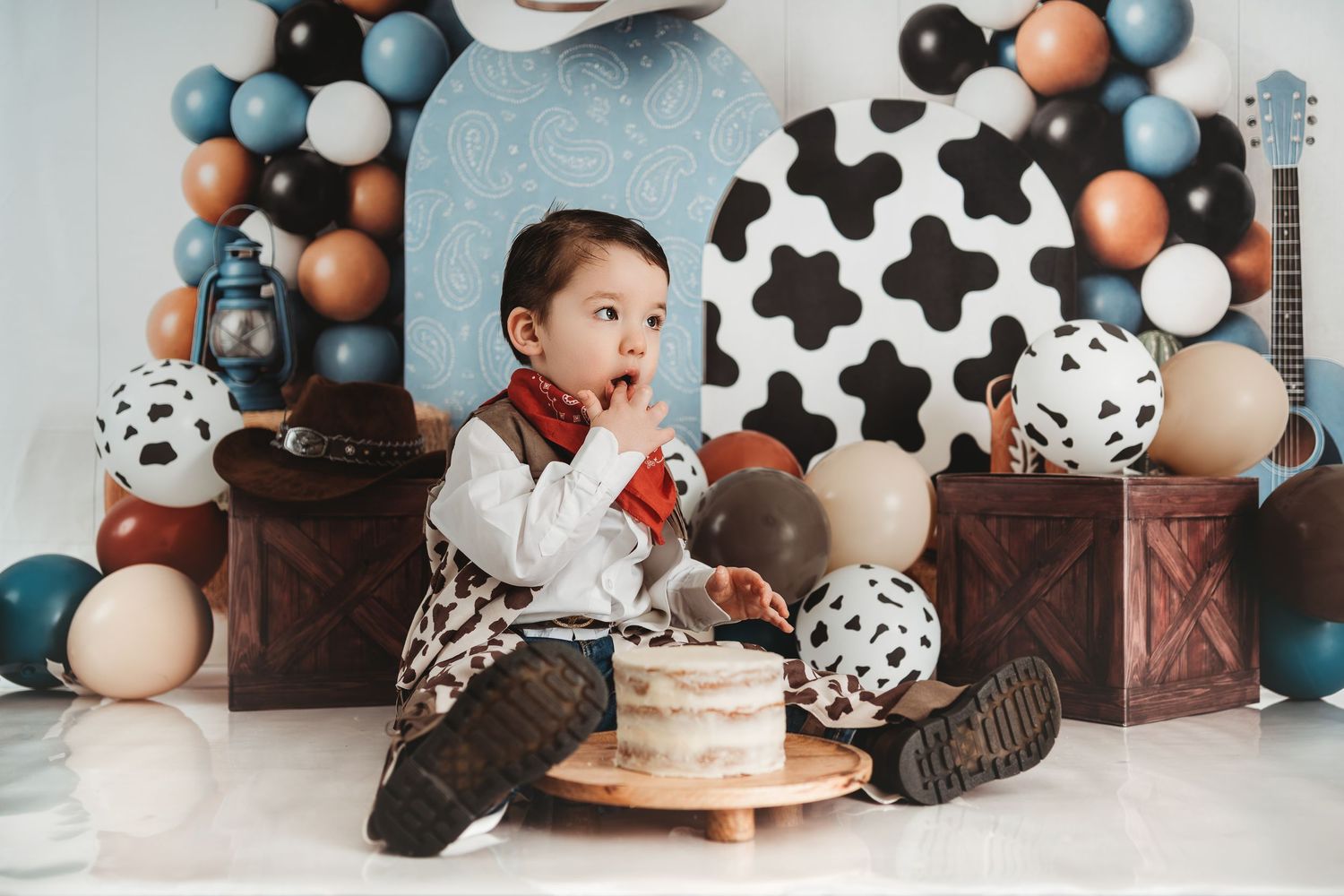 Toddler boy in cowboy outfit sits with cake smash setup, surrounded by cow print balloons and western decorations.