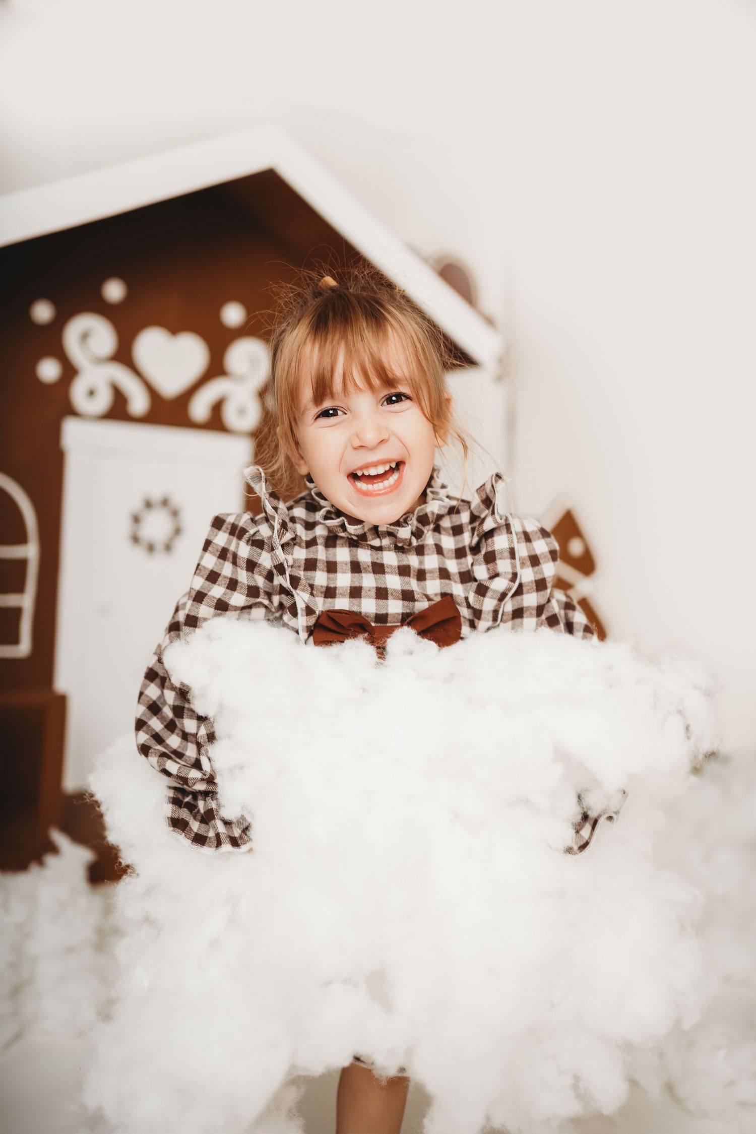 A joyful little girl in a plaid dress holds a fluffy white cloud prop in front of a decorative gingerbread house.