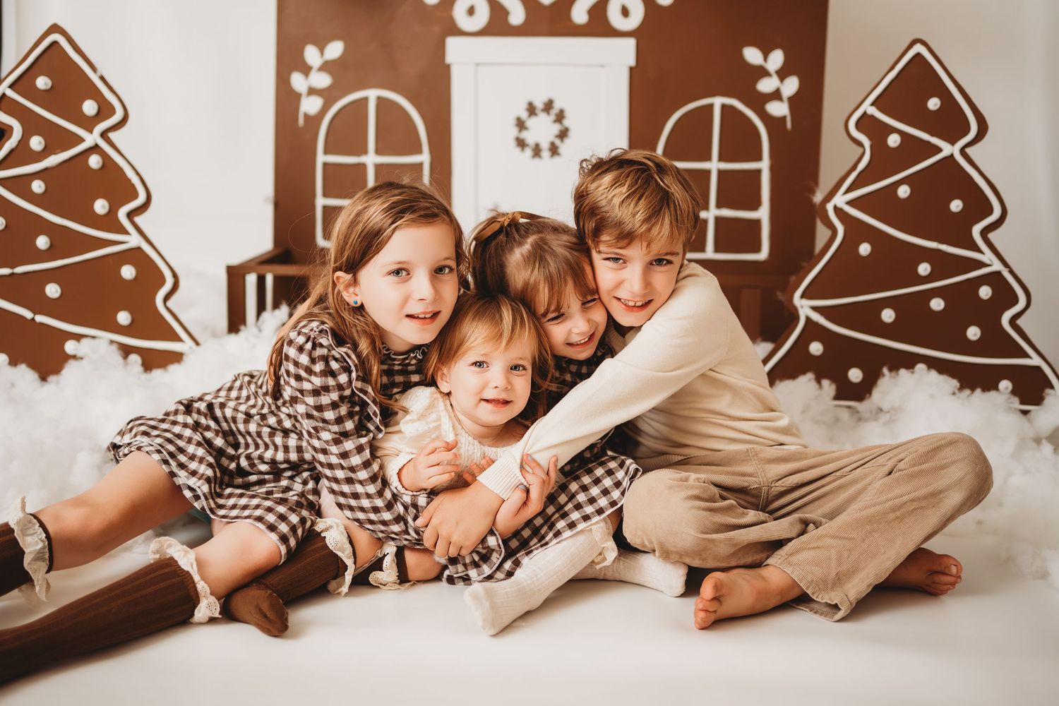 Four children hugging and smiling together in a festive Christmas gingerbread house photo studio setting.