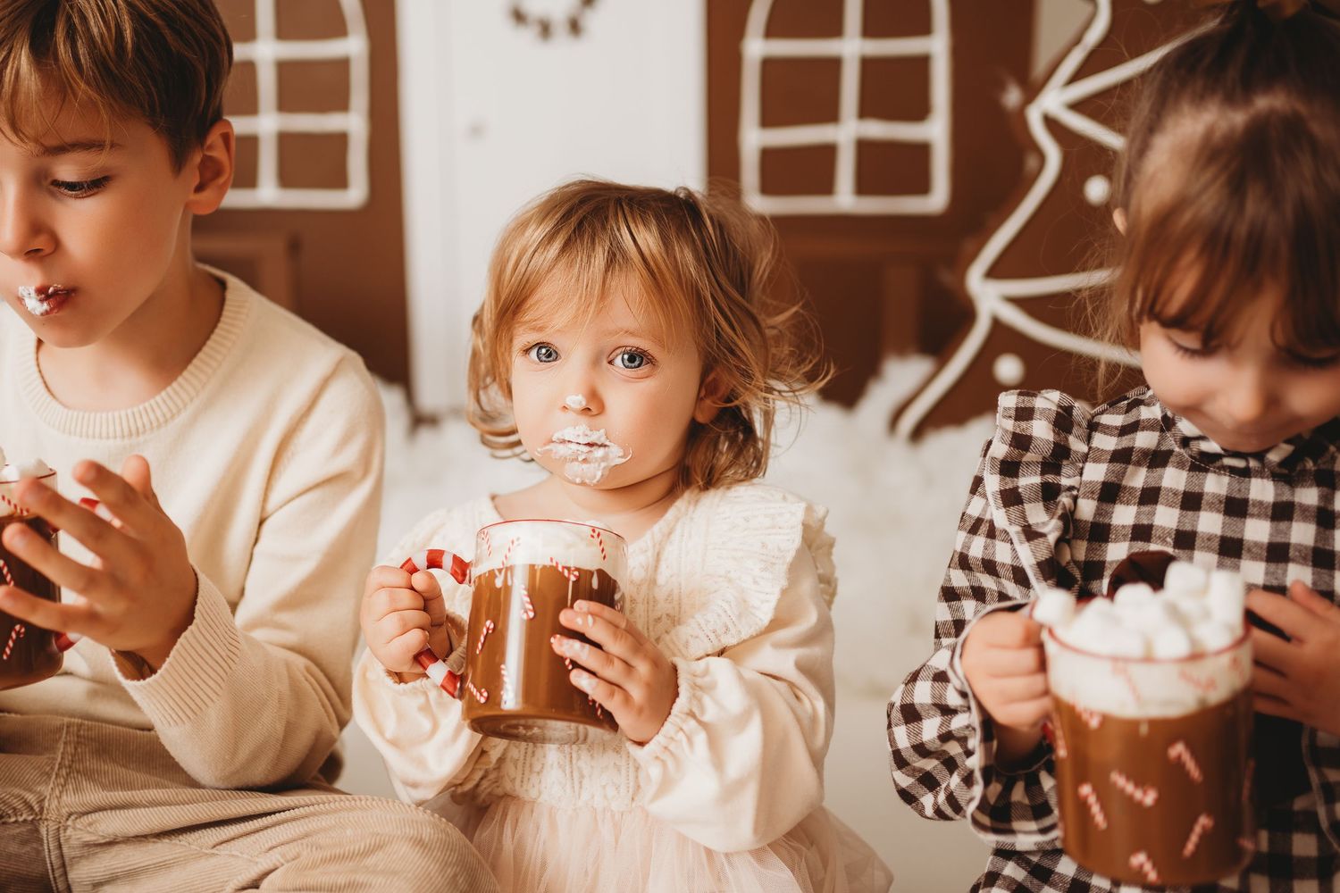 Three children sitting together drinking hot chocolate with marshmallows in front of a gingerbread house backdrop.